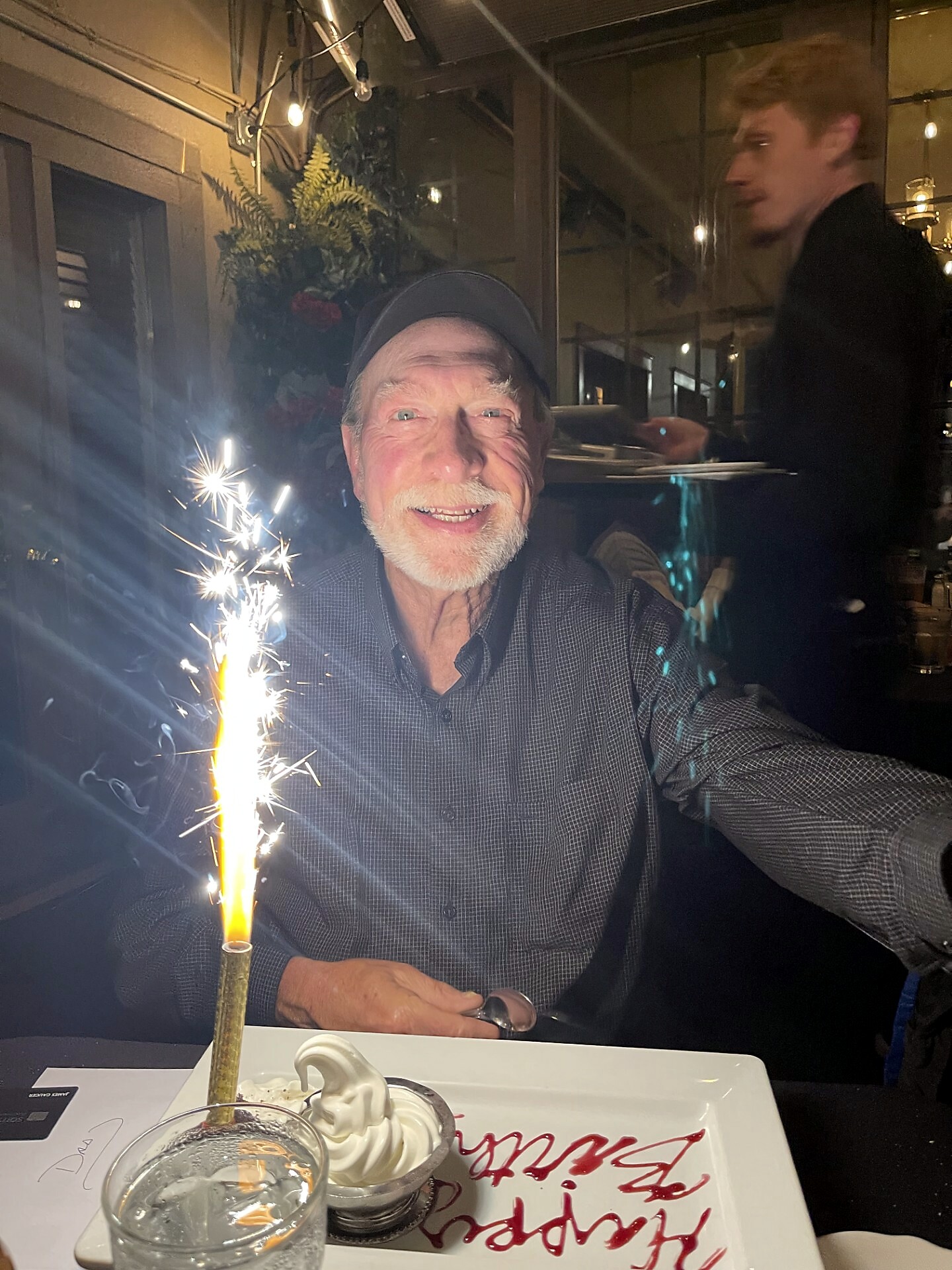 Smiling older man in a cap celebrating with a tall sparkler on a dessert at a restaurant, another person blurred in the background.