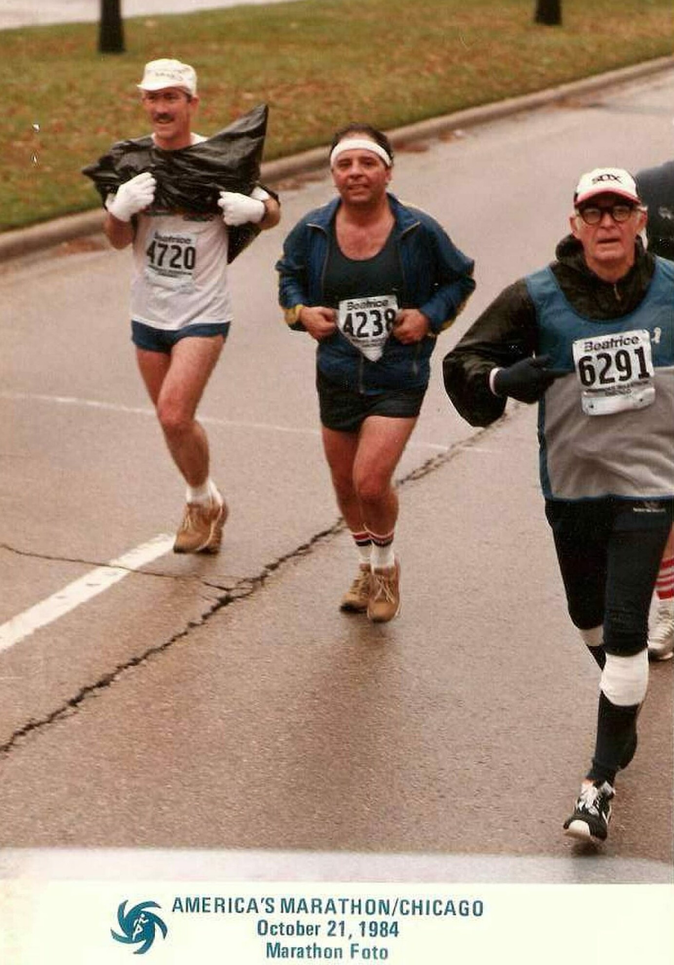 Three runners in a wet street marathon wearing bibs 4720, 4239 and 6291, mid-race on a gray day.