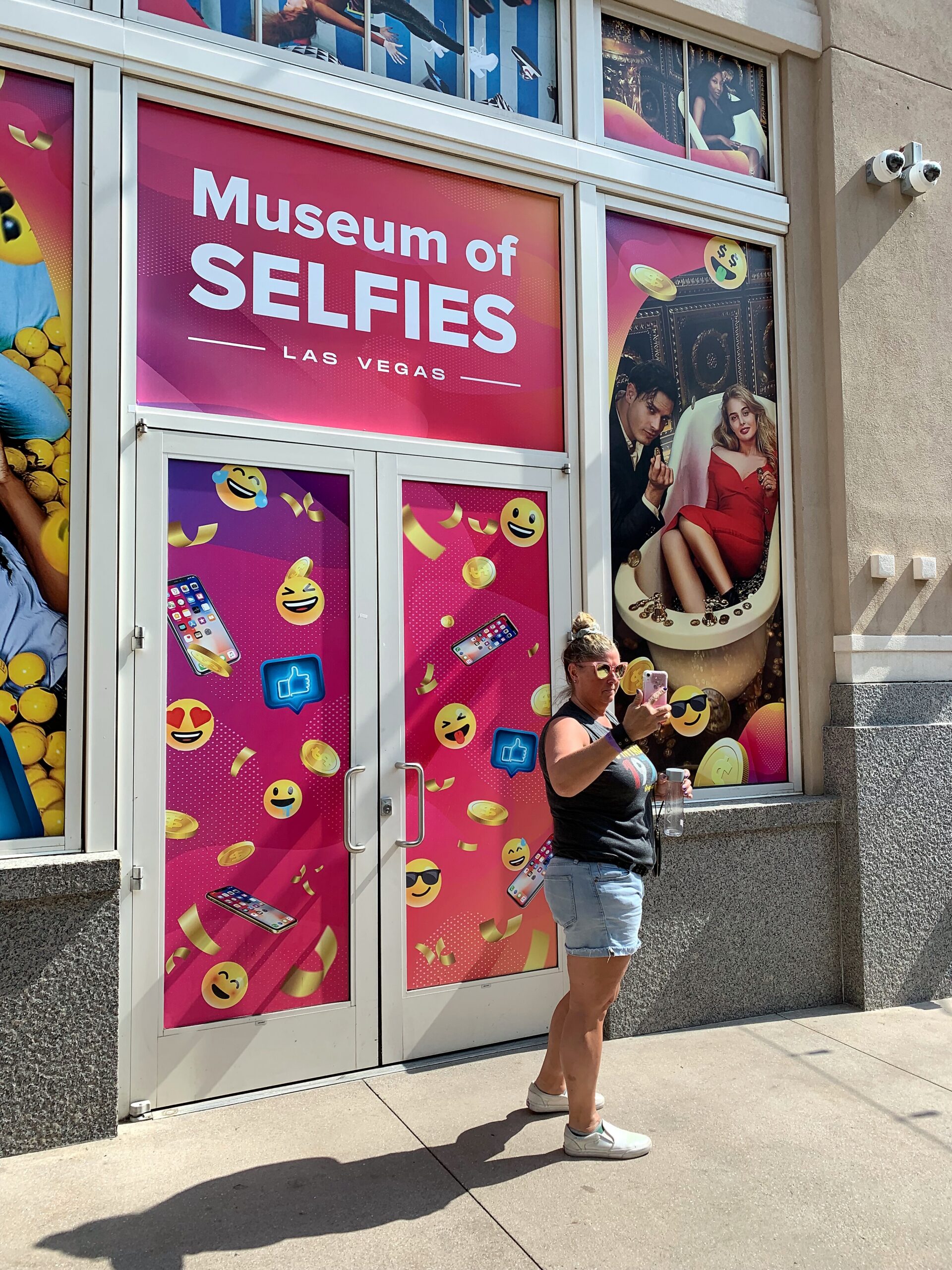 Person in a sleeveless black top and light shorts takes a selfie outside a bright pink storefront advertising the Museum of Selfies, Las Vegas.
