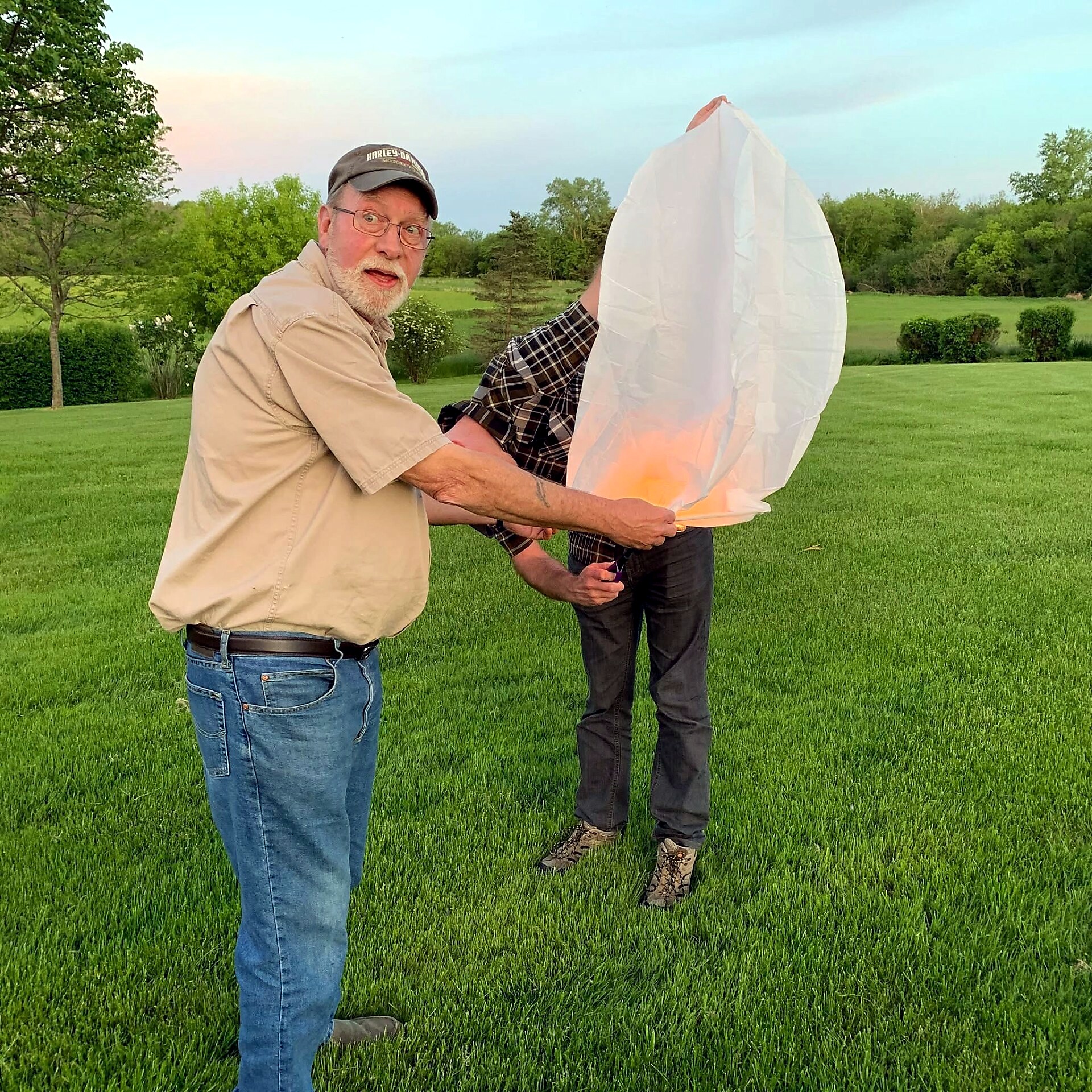 Two men outdoors on a grassy field, holding a lit sky lantern together as flames glow inside the paper lantern in the evening light.