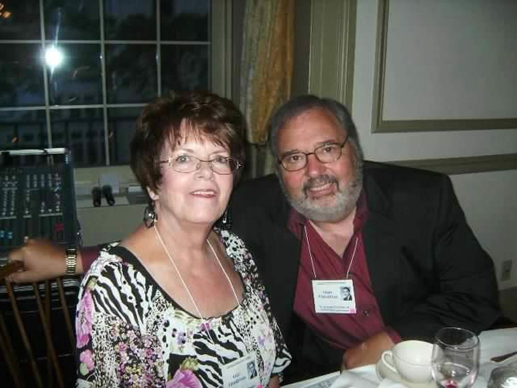 Smiling woman and man pose at a table during a social event, both wearing name badges on lanyards.