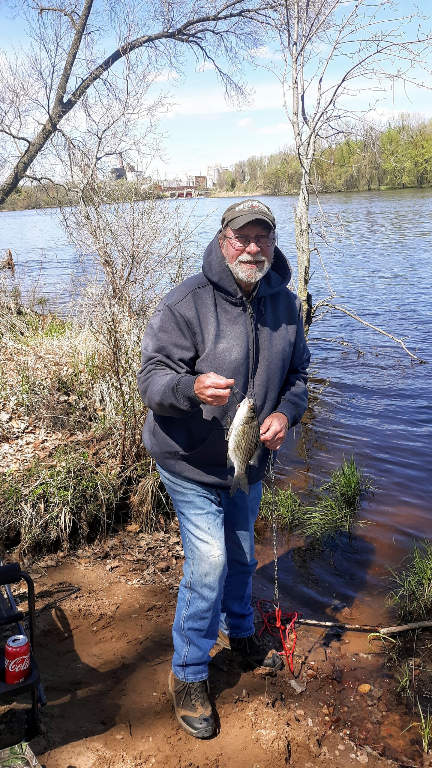 Older man in a hoodie and cap holding a fish by a riverside, with trees and a distant city skyline in the background.