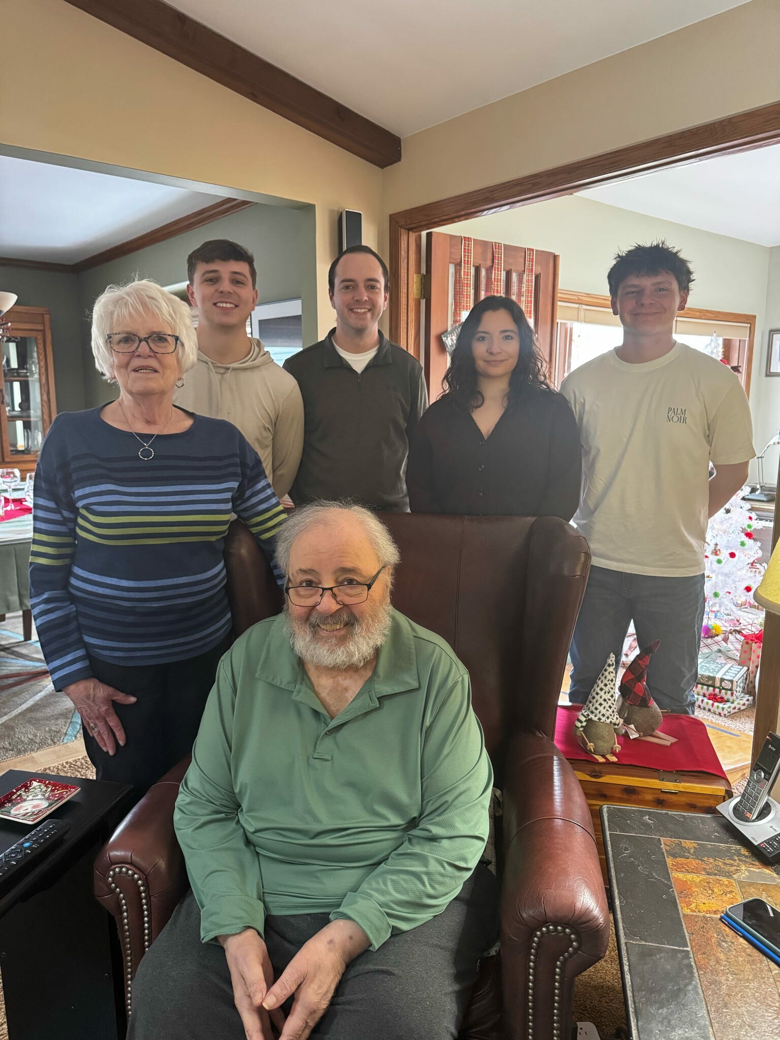 Elderly man with glasses seated in a leather chair in a living room, with five relatives standing behind him and a Christmas tree in view.
