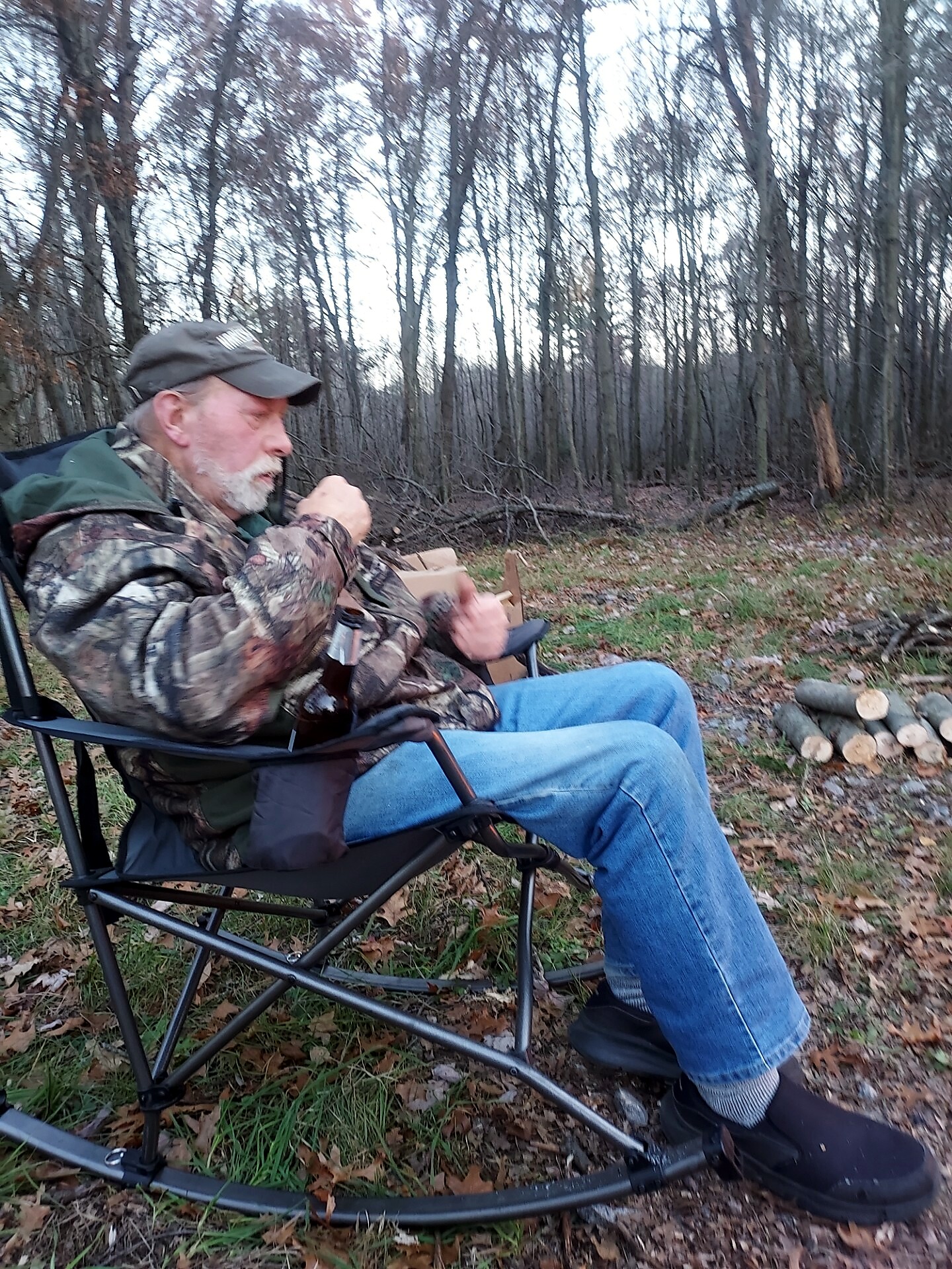 Older man with a gray beard wearing a camo jacket and cap sits in a folding camping chair in a forest, nearby stacked logs ready for firewood.