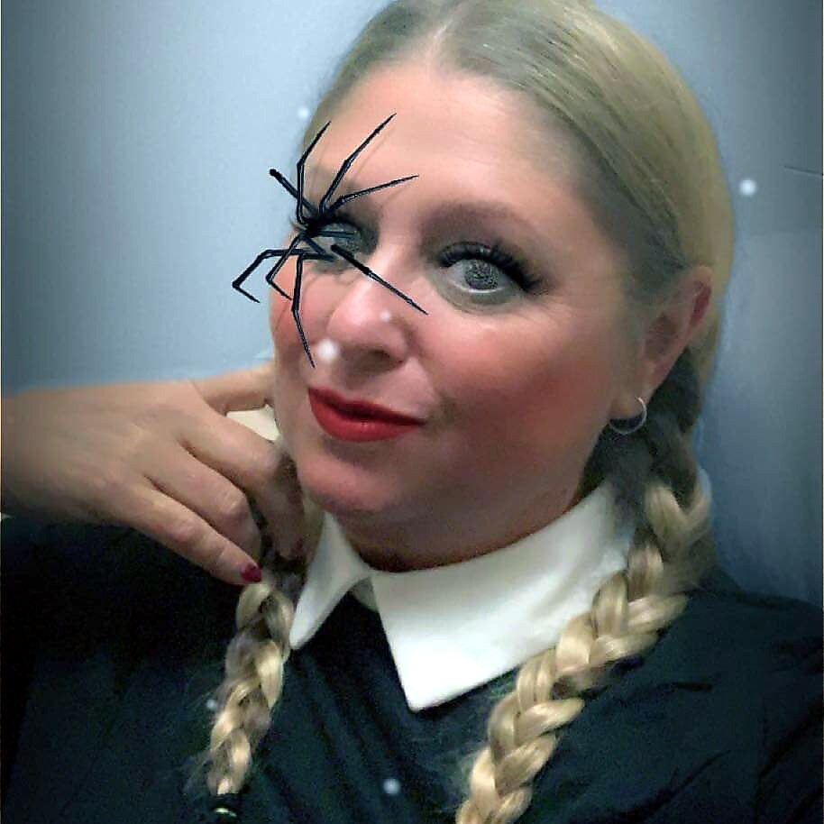 Woman with blonde braids and red lipstick has a large fake spider perched on her left eye area, smiling at the camera.