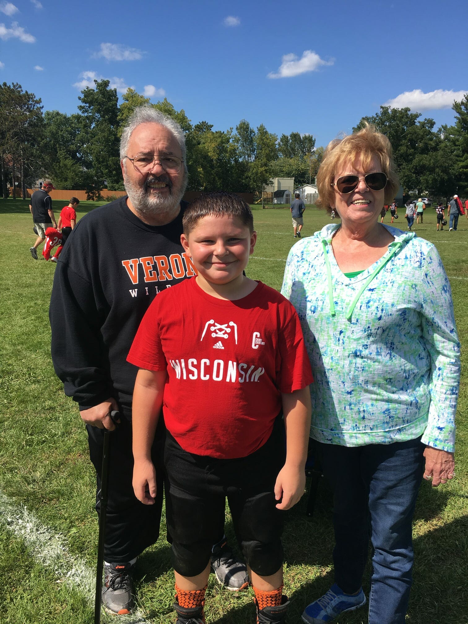 A smiling trio outdoors on a grassy field: an older man with glasses and a beard, a boy in a red Wisconsin shirt, and a woman in sunglasses standing together.