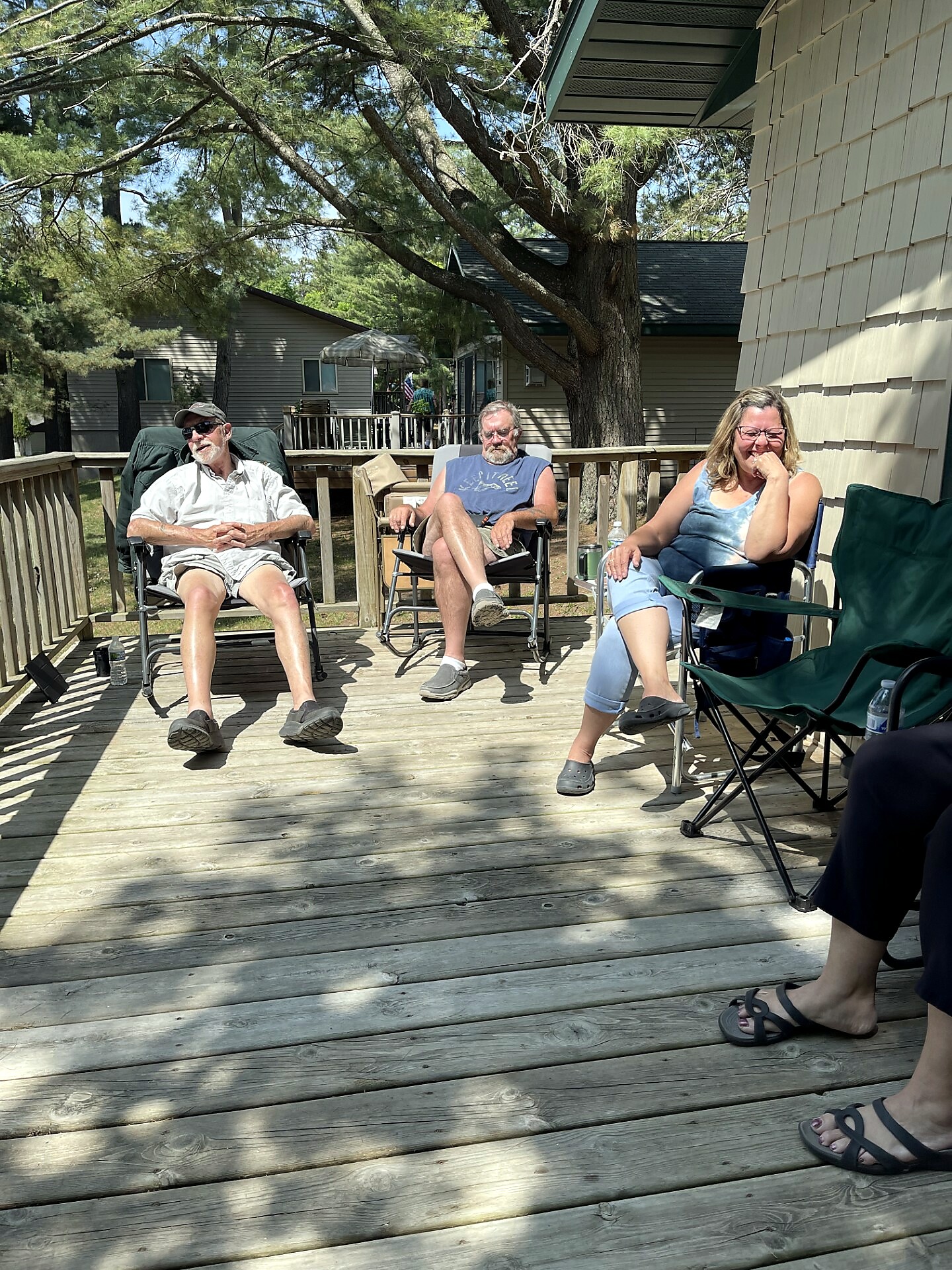 Three adults sit in folding chairs on a wooden deck outside, with trees and houses in the background; they appear relaxed and casually dressed.