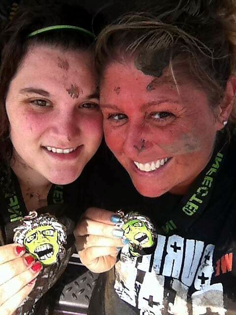 Two smiling women with muddy faces posing closely, each holding a zombie-themed medal on a ribbon.