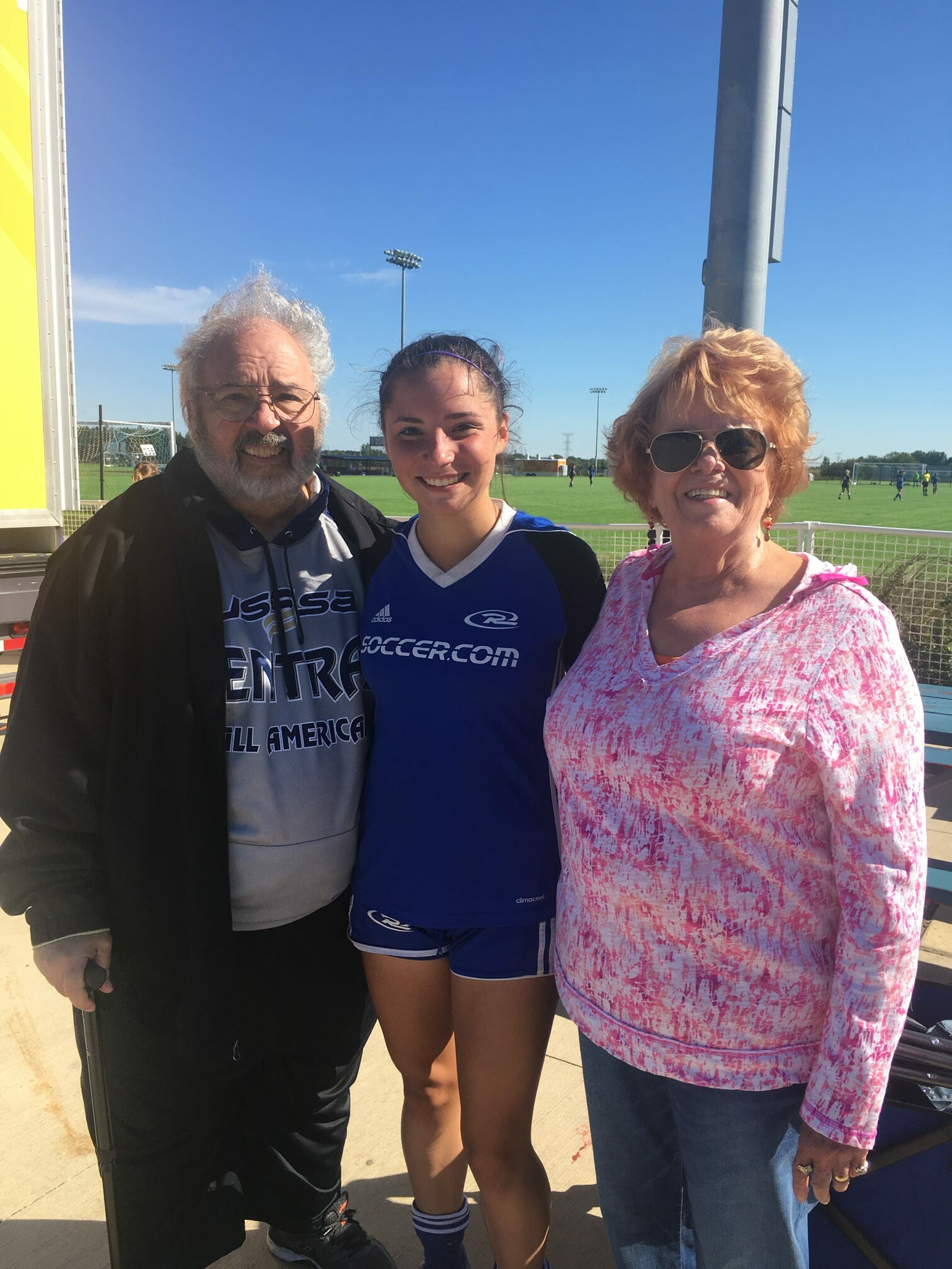 Three smiling people pose together at a sunny soccer field; older man left, young player center in blue jersey, older woman right in pink tie-dye top and sunglasses; goal and field in background.