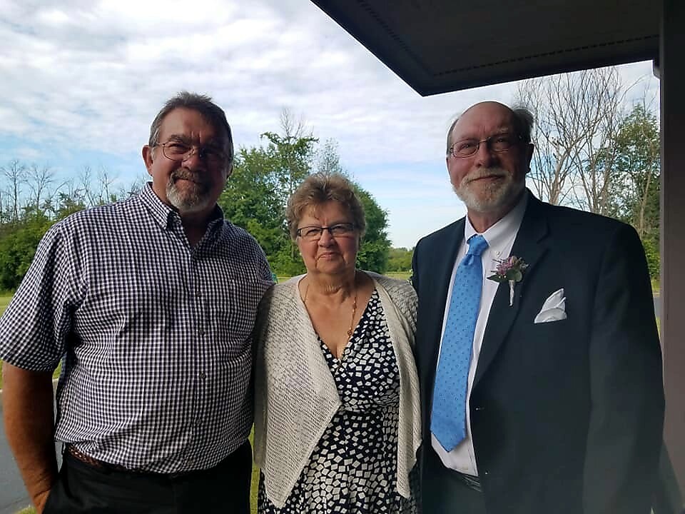 Three adults posing together outdoors on a sunny day; a man in a suit with a blue tie and boutonniere stands on the right, a woman in a patterned dress and cardigan in the middle, and a man in a checkered shirt on the left.