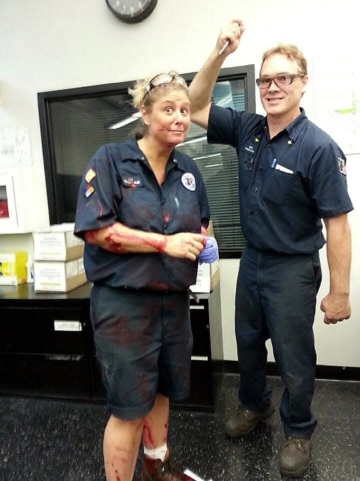 Two people in navy work uniforms pose in a clinical room; the woman has fake blood on her arms and leg wrap, smiling as the man holds up a small object.