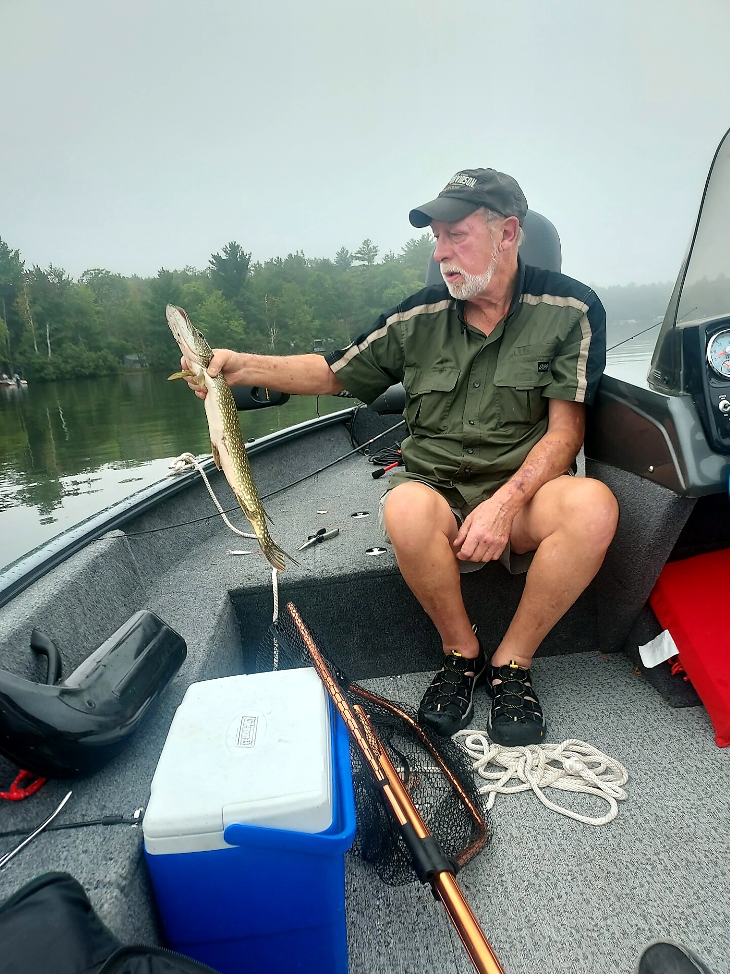Older man on a boat holding a long fish by the mouth, with a lake and forest in the background.