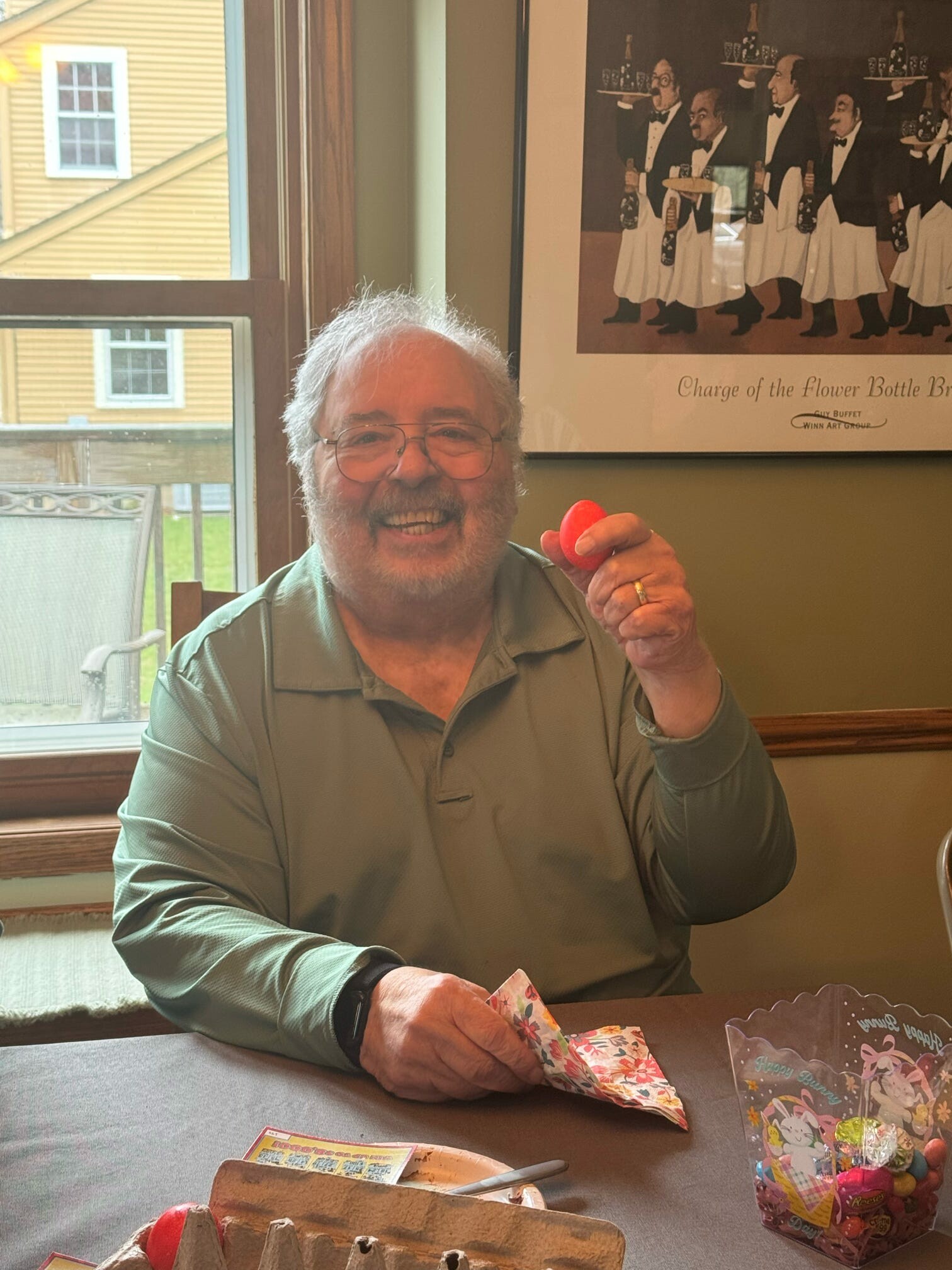 Smiling older man with glasses holds a red plastic Easter egg at a table with candy and crafts in a sunlit room.