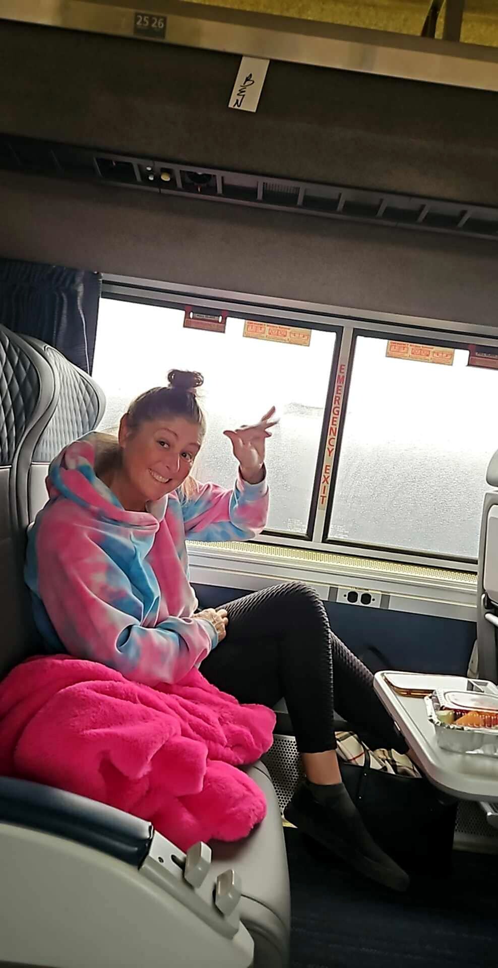 Young woman in a pastel tie-dye hoodie sits on a bus seat with a bright pink blanket, smiling and making a peace sign toward the camera.