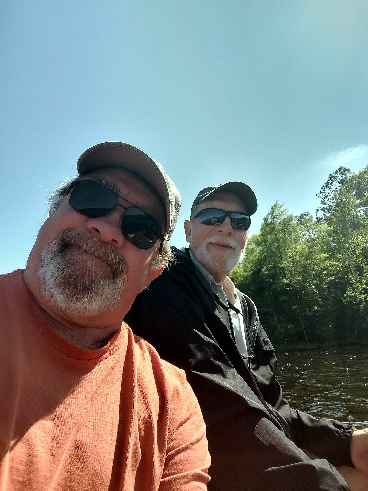 Two men wearing hats and sunglasses sit in a boat on a sunny day with water and trees in the background (selfie).