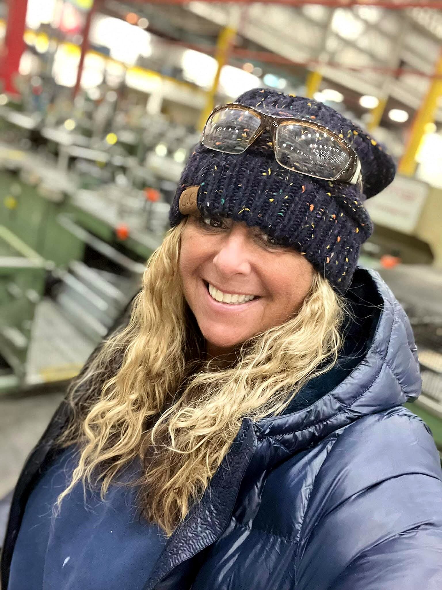 Smiling woman wearing a dark knit hat with colorful flecks and sunglasses on top, in a blue puffer jacket in an indoor shop or workshop.