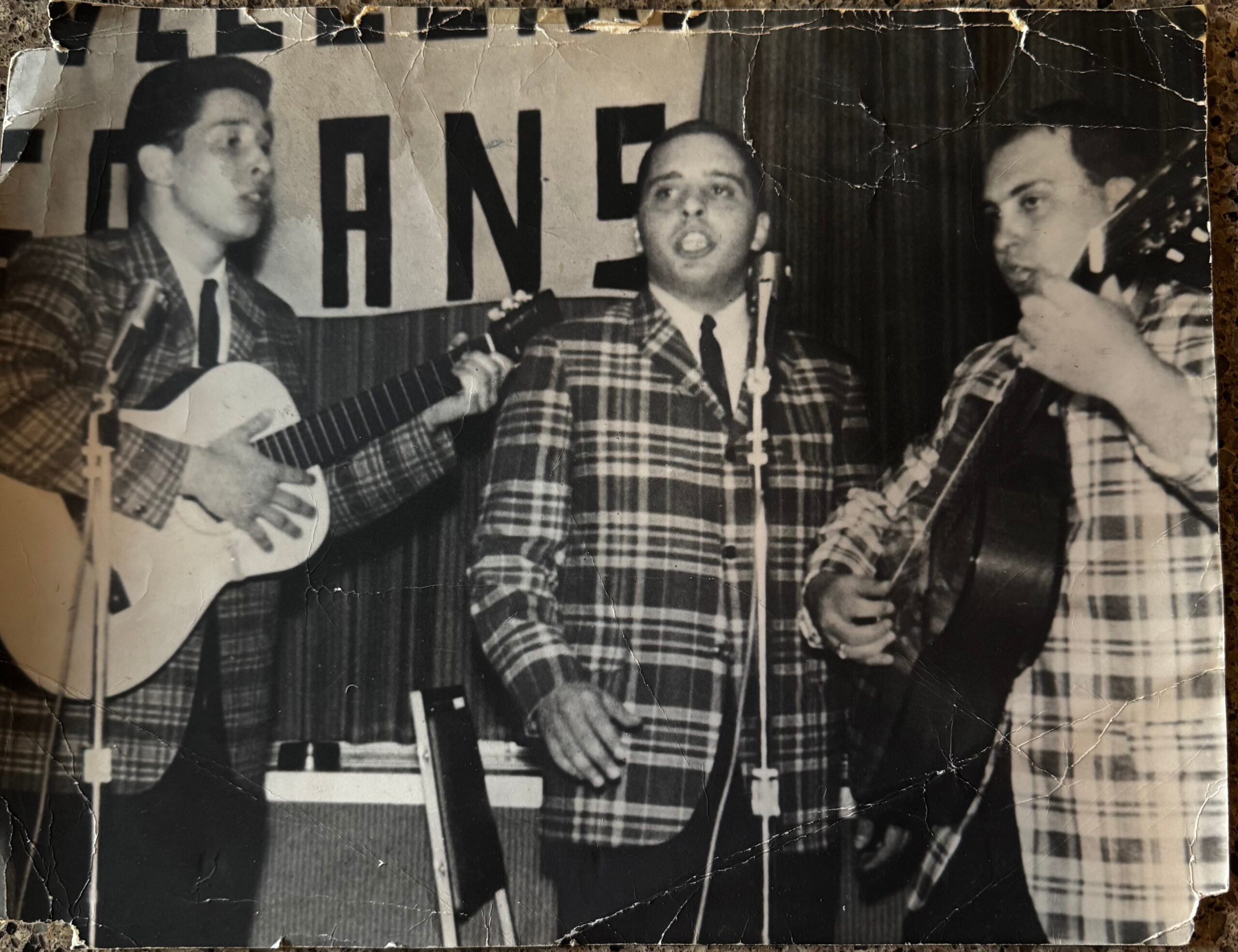 Three men in plaid suits performing together on stage, with guitars and a microphone, likely a vintage vocal group.
