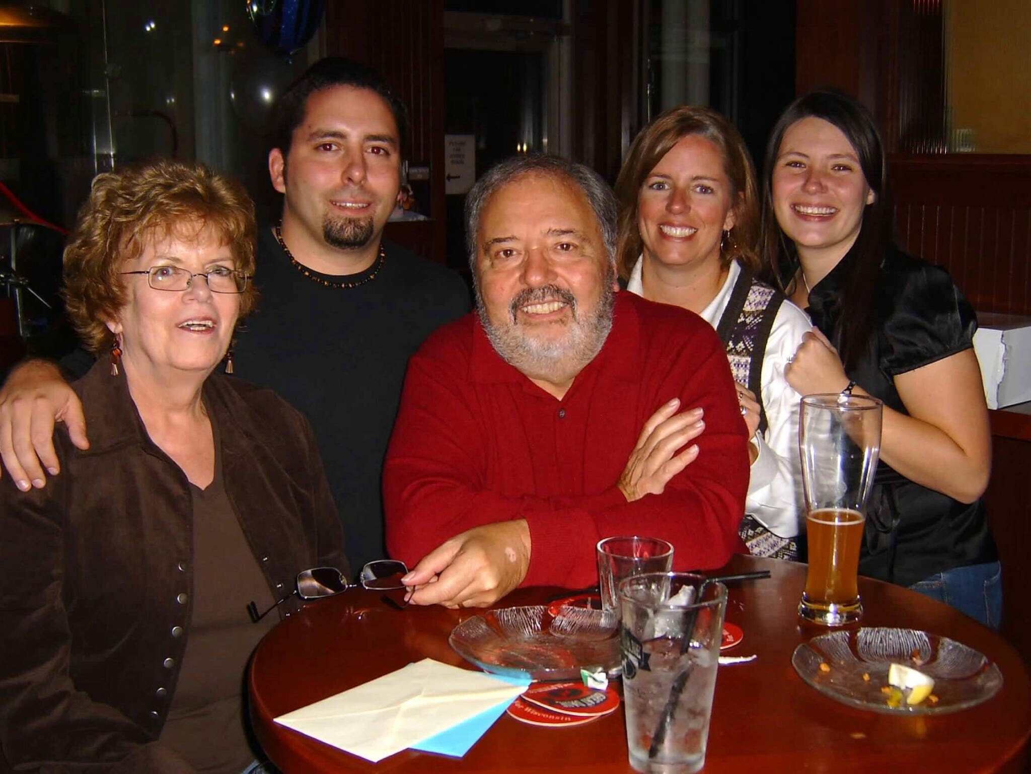 Five adults smile for a group photo around a restaurant table, with drinks and napkins visible on the table and balloons in the background.
