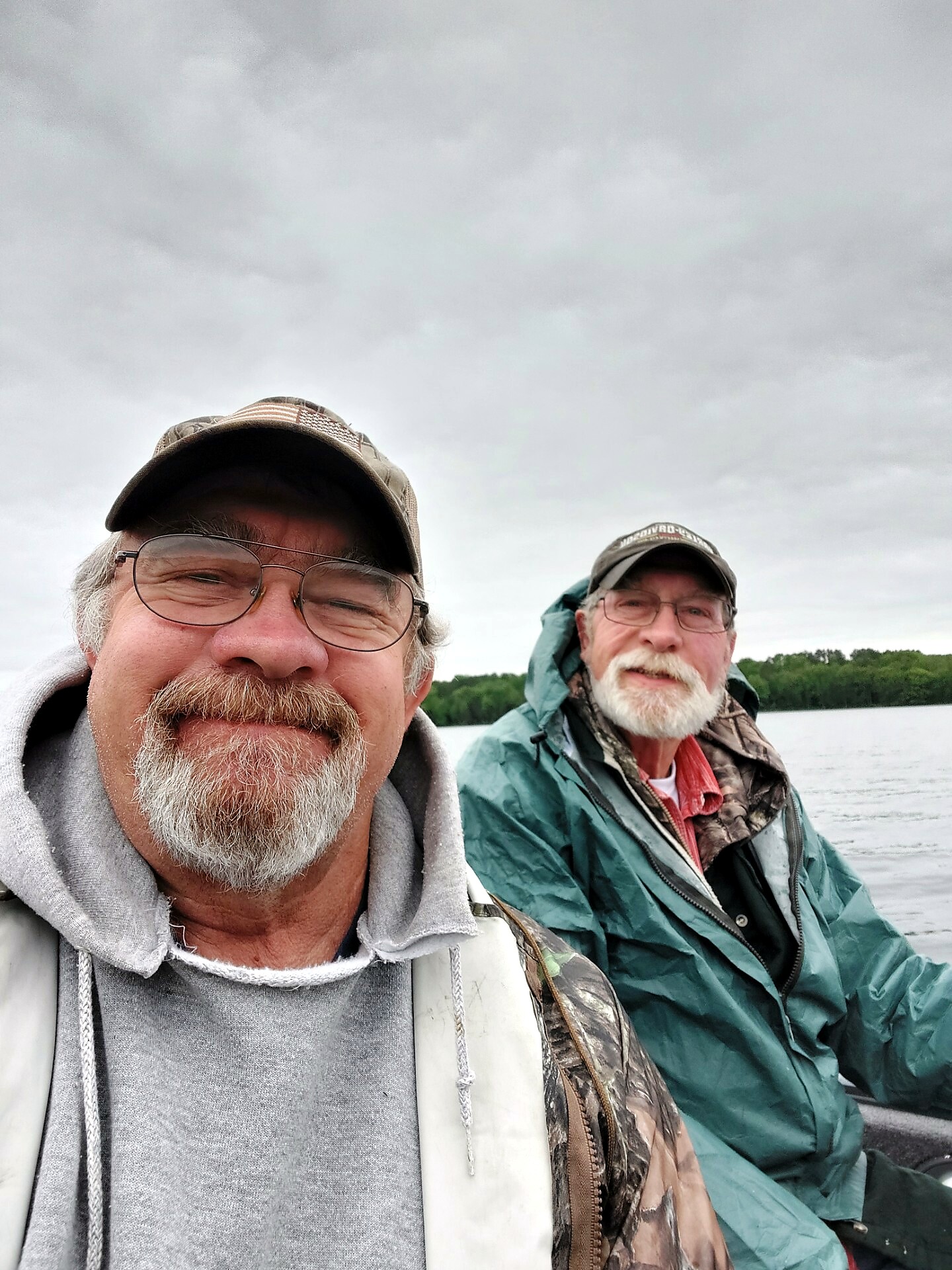 Two men wearing caps and jackets on a boat, with a gray cloudy sky and lake in the background.