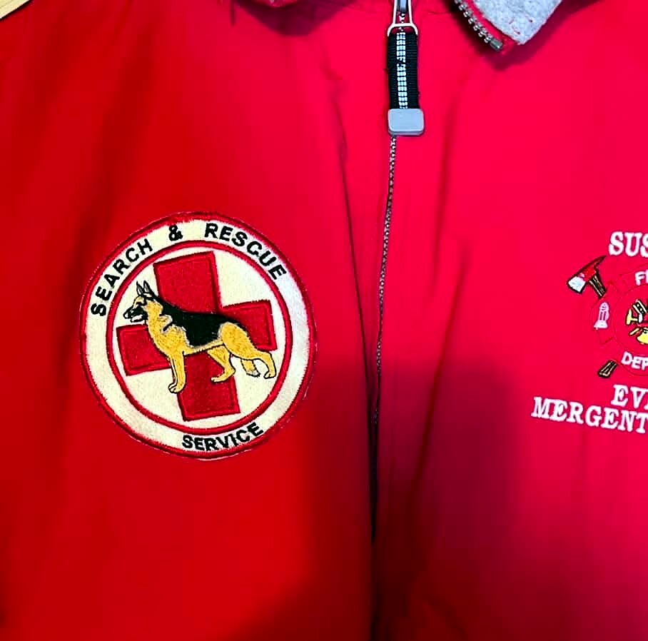 Close-up of a red rescue jacket with a circular 'Search & Rescue Service' patch showing a German Shepherd in front of a red cross.