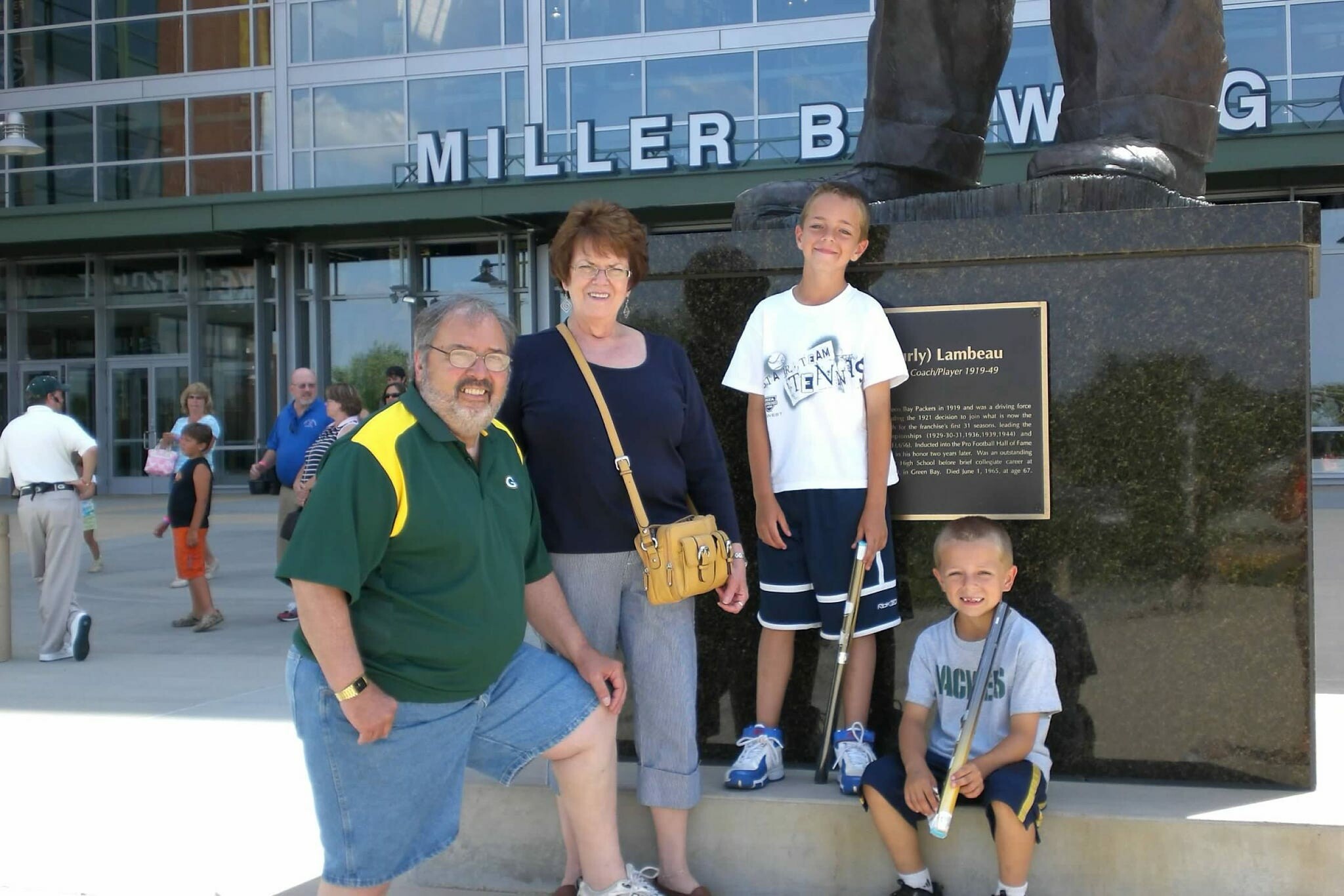 Family posing in front of a large statue at Miller Brewing Co. exterior, with a plaque and glass-front building in the background.