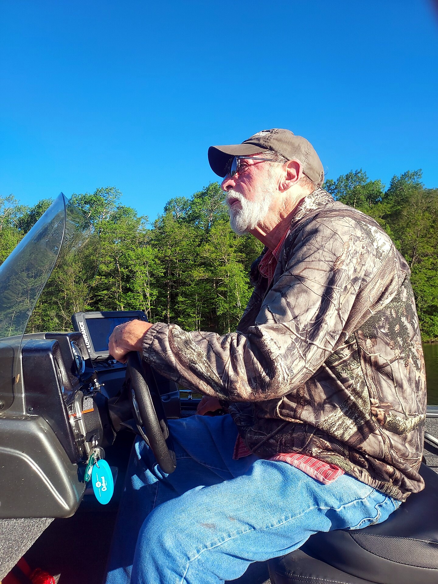 Older man with a white beard driving a camo jacketed boat, wearing cap and sunglasses, steering at dashboard against blue sky and trees in background