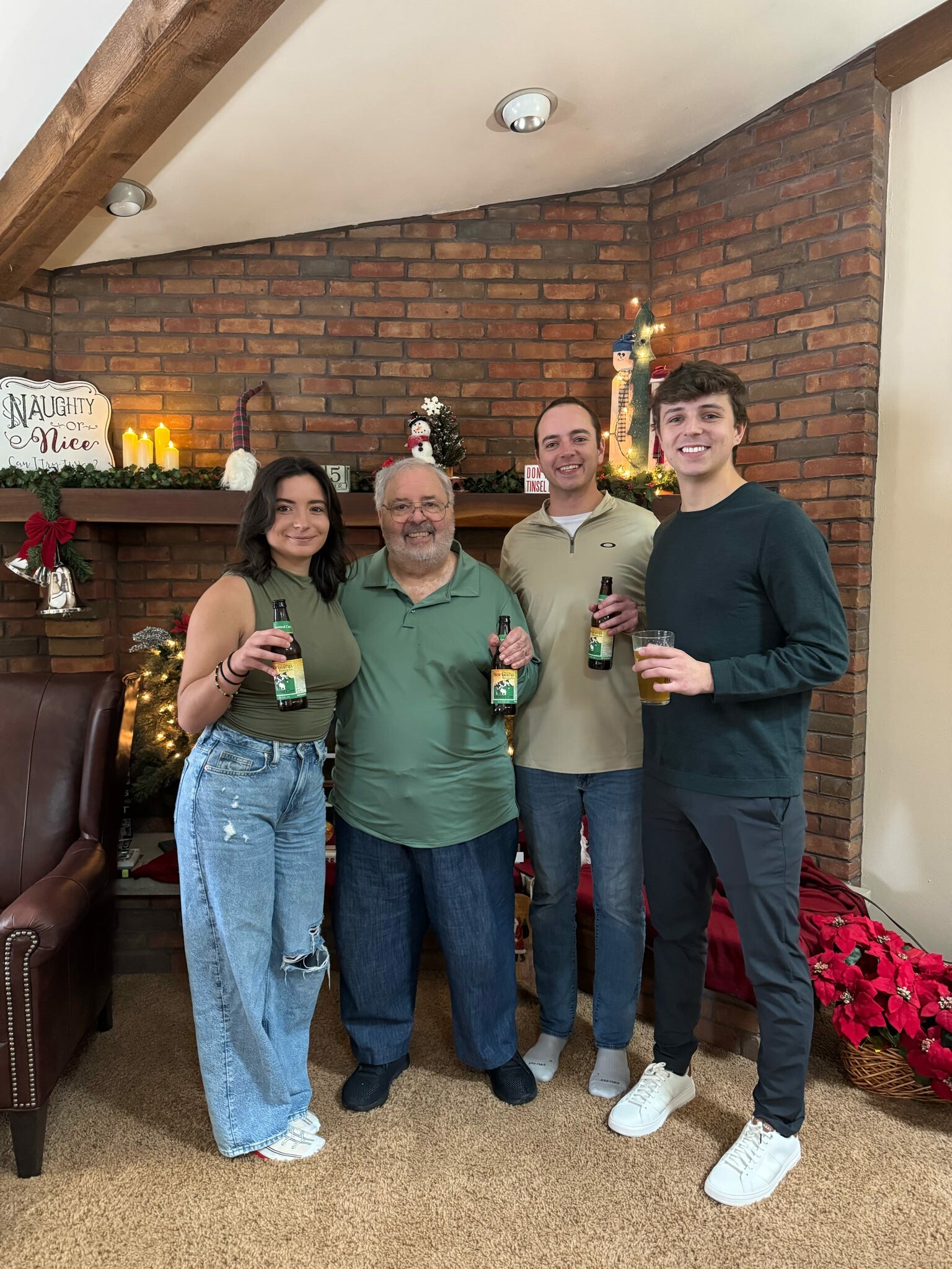 Four people standing together indoors holding beer bottles in a Christmas-decorated living room by a brick fireplace.