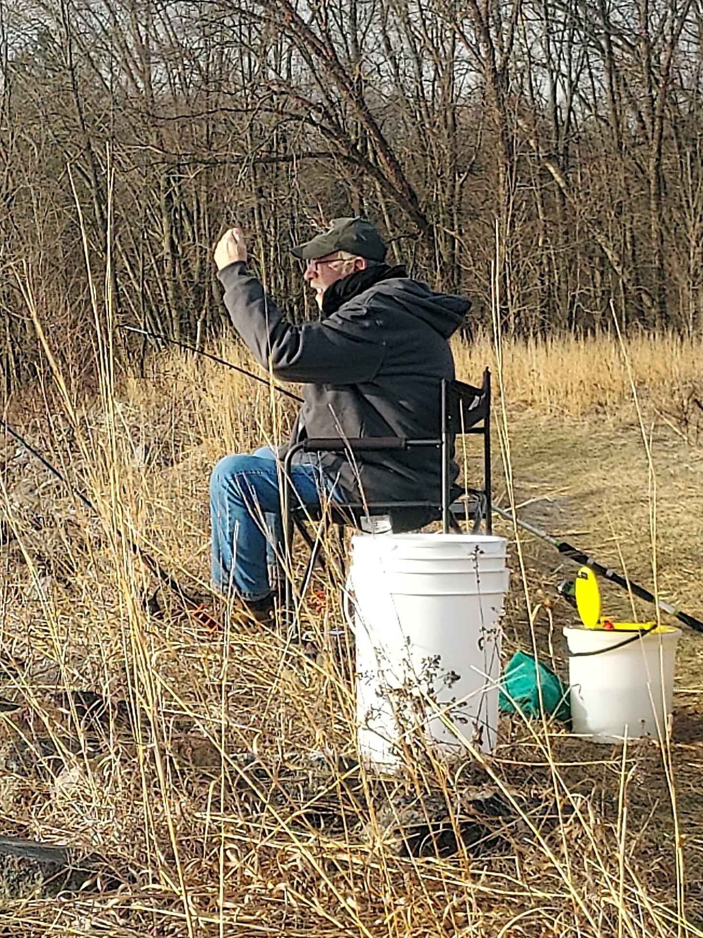 Man fishing from a foldable chair in a marshy, grass-filled area, wearing a dark jacket and cap with a fishing rod in hand and a white bucket nearby.