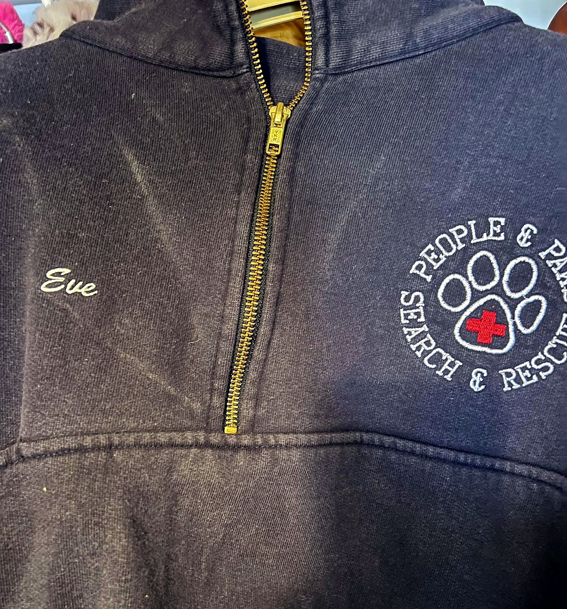 Close-up of a dark navy zip-up jacket showing a brass zipper, with partial white embroidery and a circular emblem containing a red cross on the right.