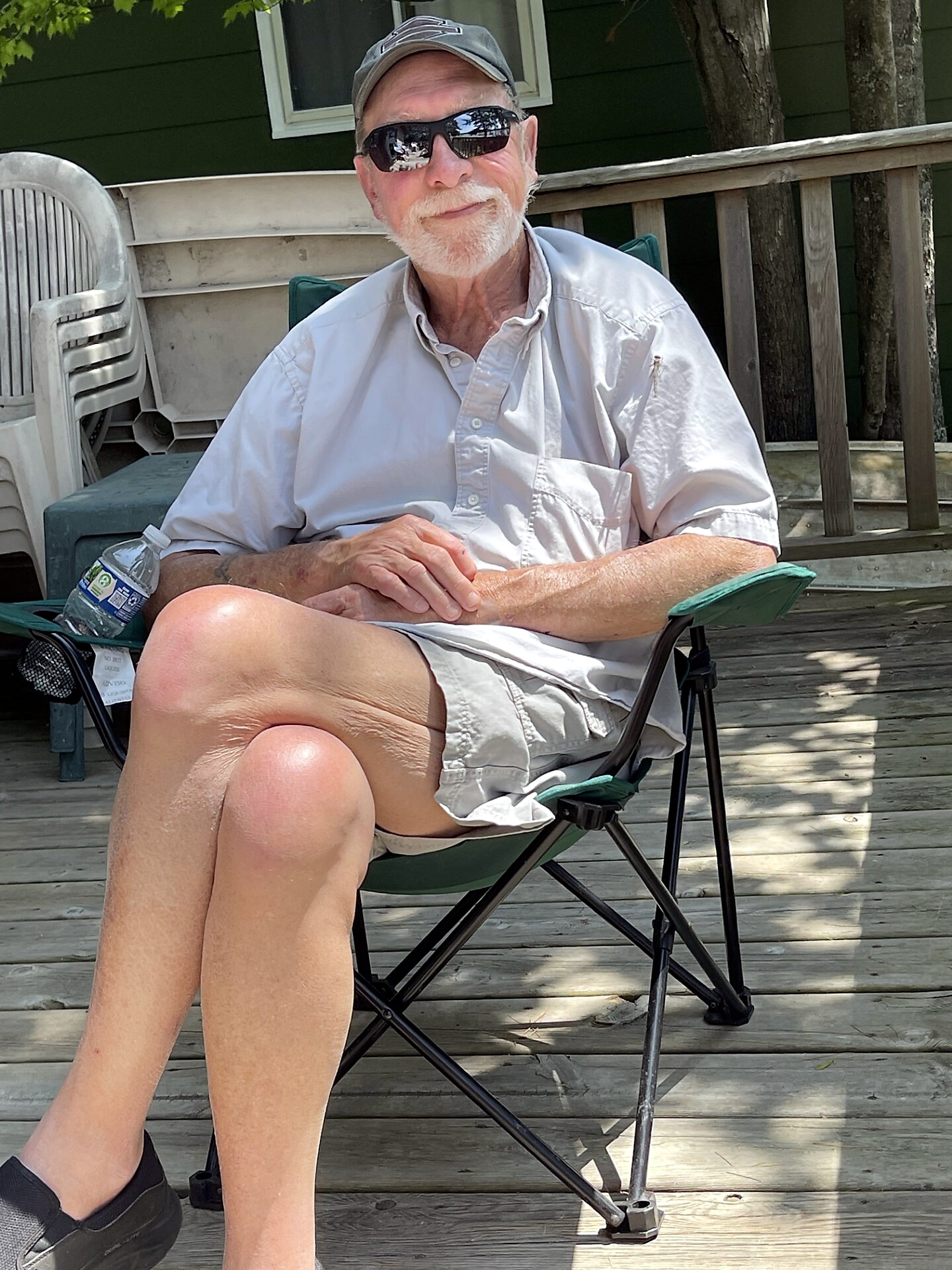 Older man with a white beard sits on a green folding chair on a wooden deck, wearing a cap, sunglasses, light shirt and shorts.