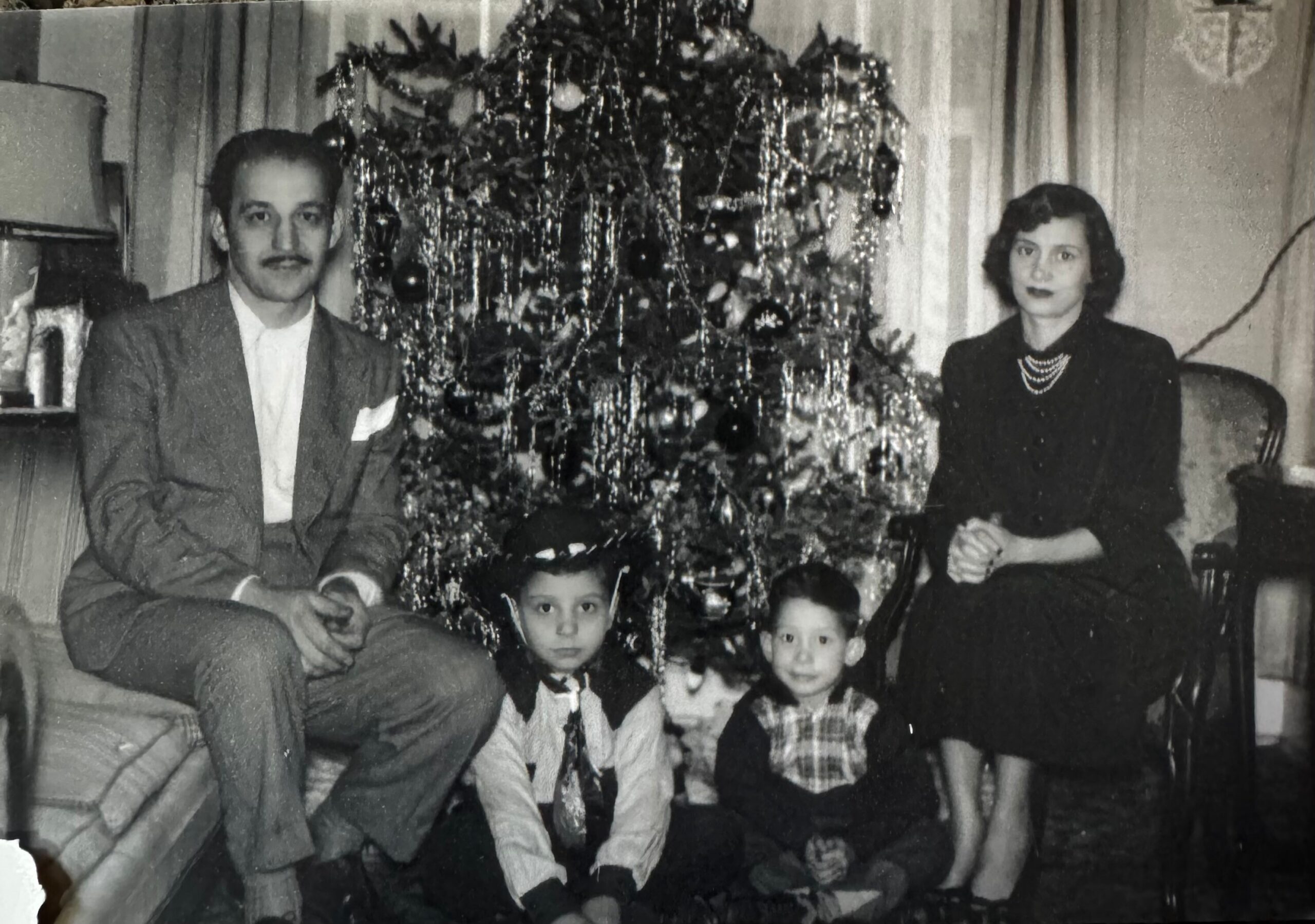 Family of five seated and standing around a Christmas tree in a living room, posing for a holiday photo.