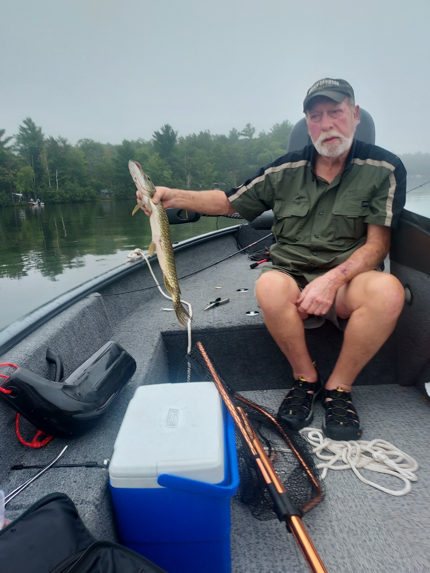 Older man in a green shirt and cap sits in a boat on a calm lake, holding up a long northern pike he just caught.