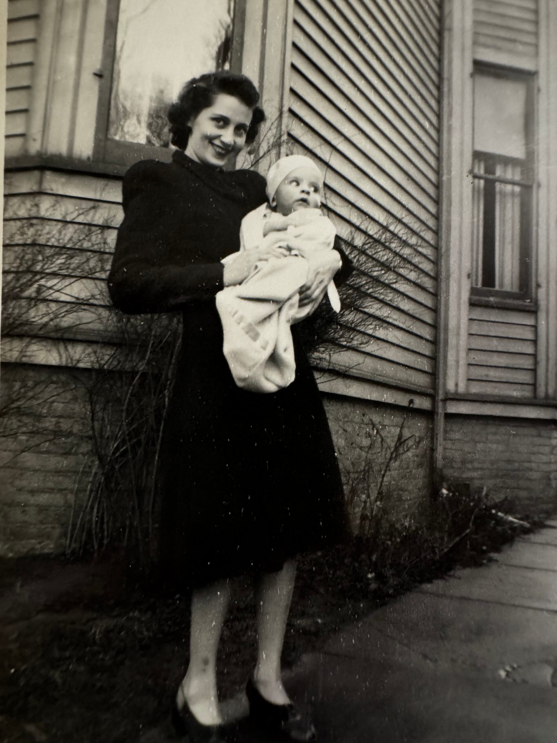 A smiling woman in a dark coat holds a baby wrapped in a light blanket outside a wooden house.