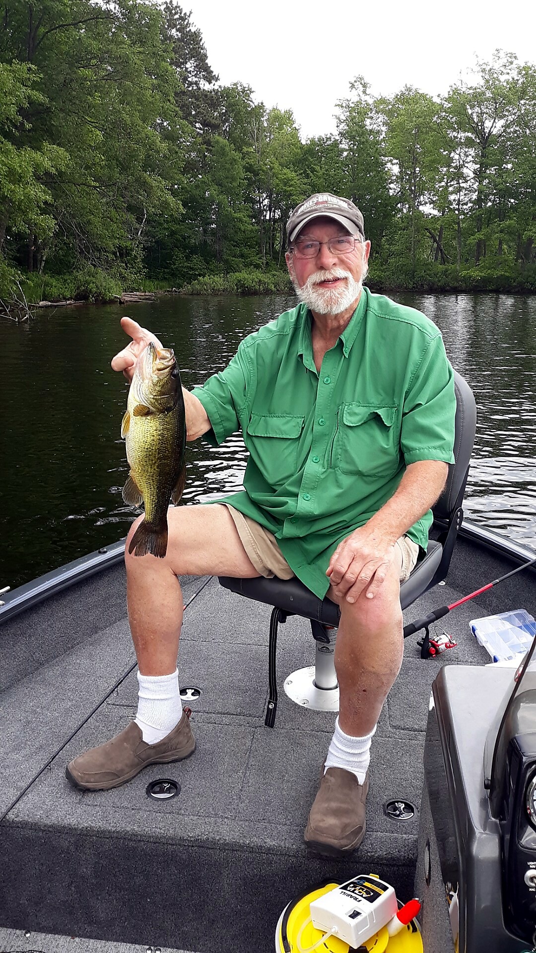 Older man with a white beard sitting on a boat, holding up a largemouth bass he caught.