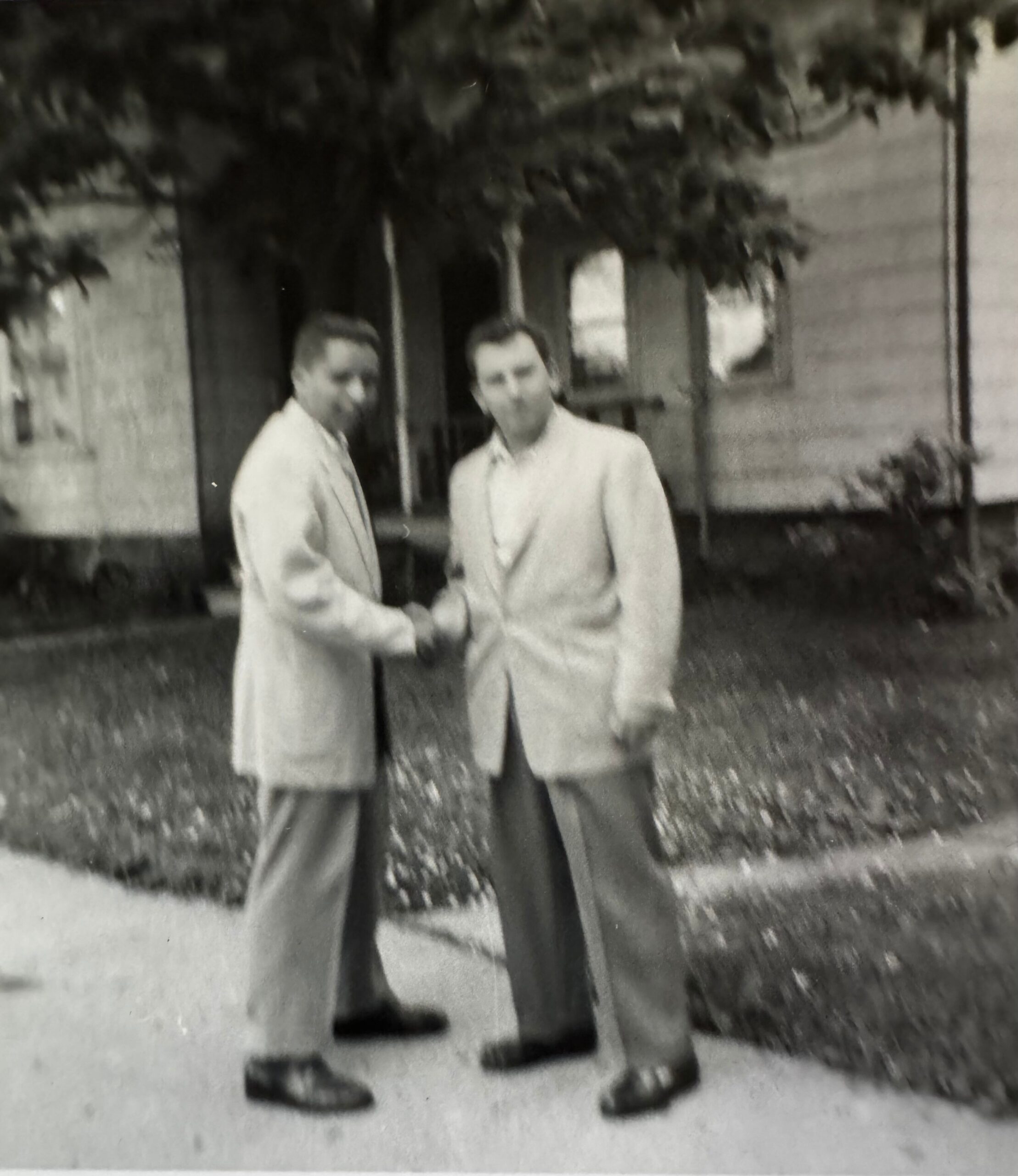Two men in light suits shake hands outside a building with arched doorway, trees in the background (old, black-and-white photo).