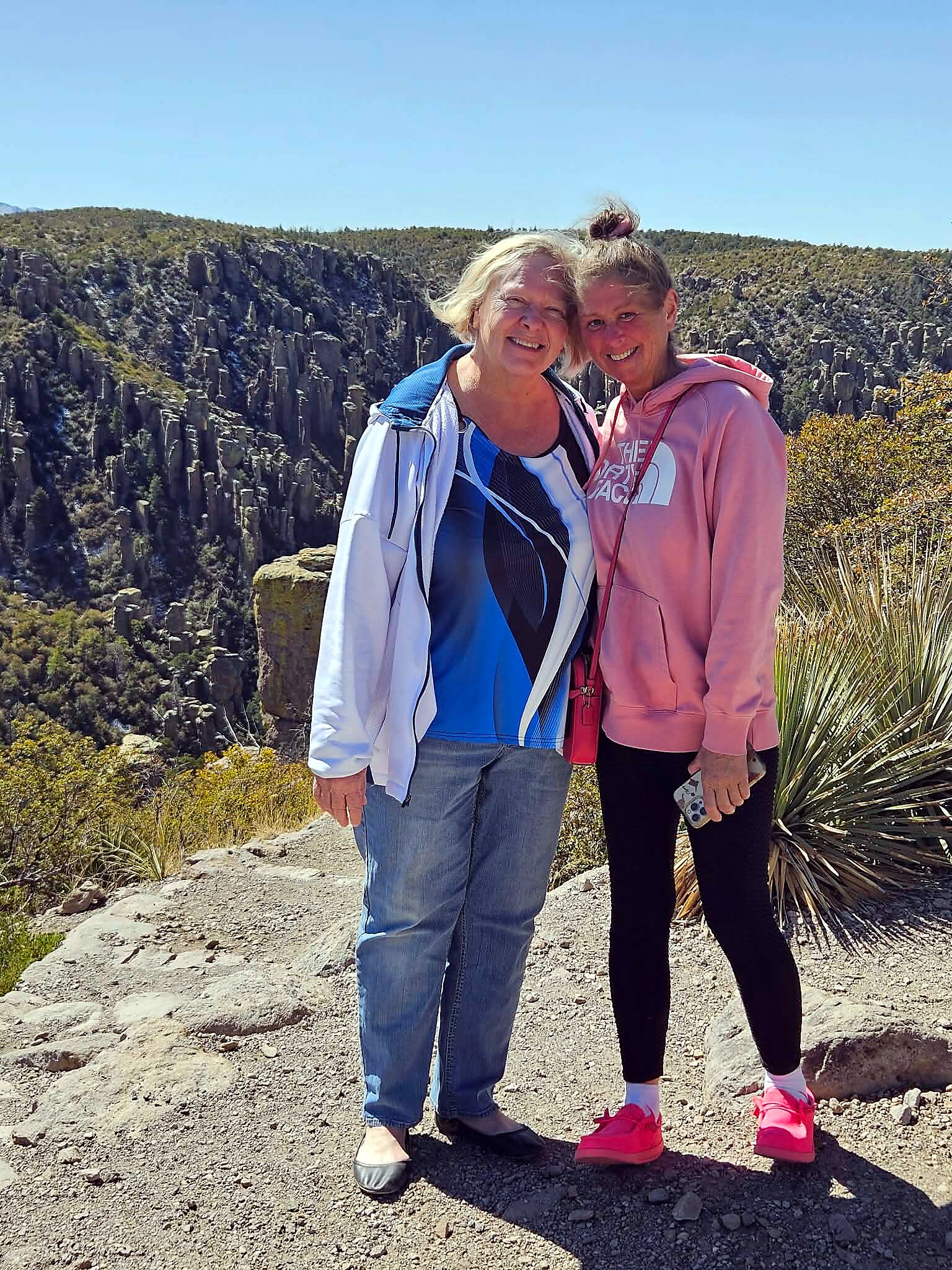 Two women stand together on a rocky overlook, smiling with a rugged canyon landscape behind them on a sunny day.