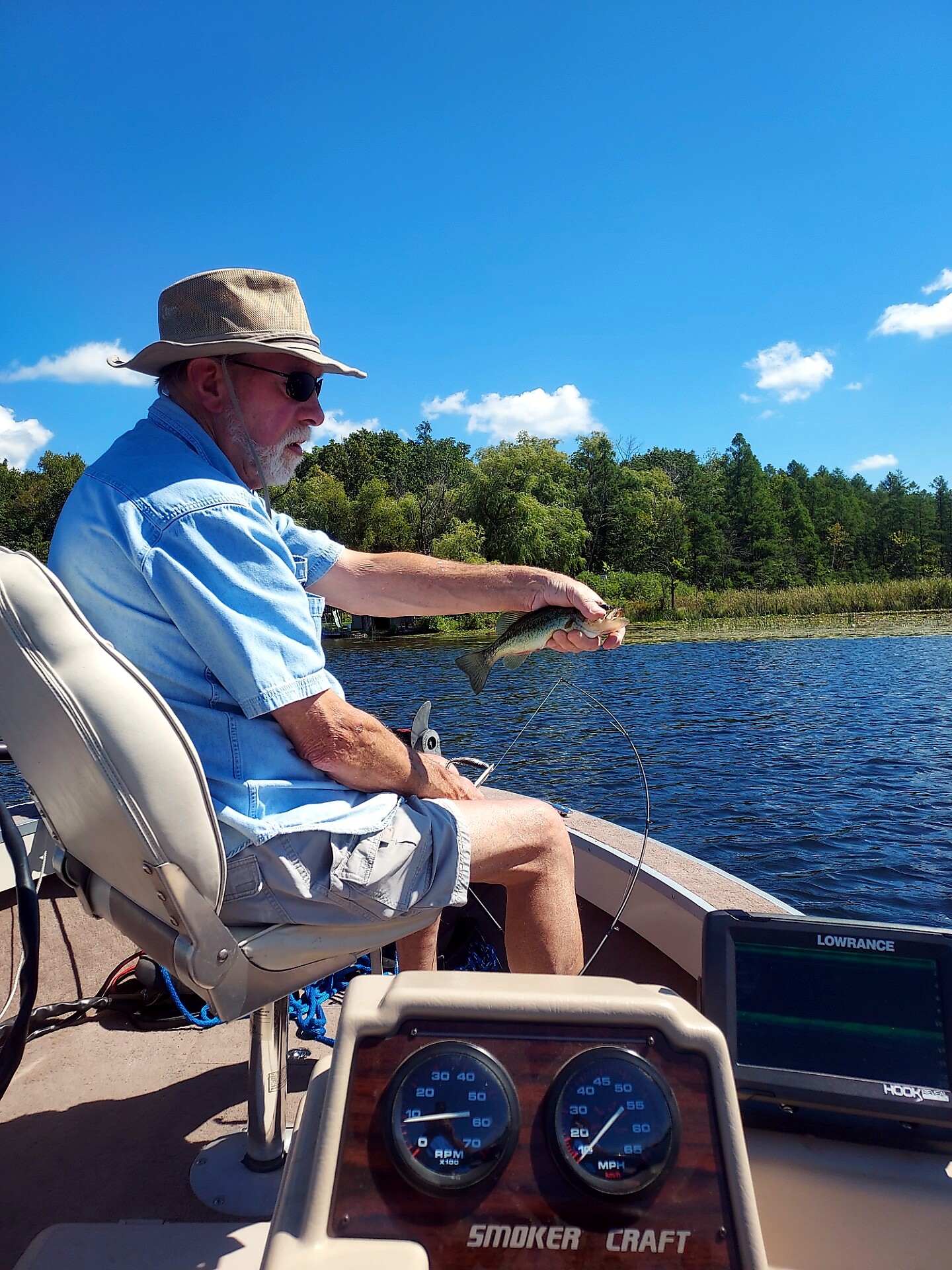 Older man in a hat and sunglasses sitting on a boat, holding a small fish over calm lake with trees in the background.