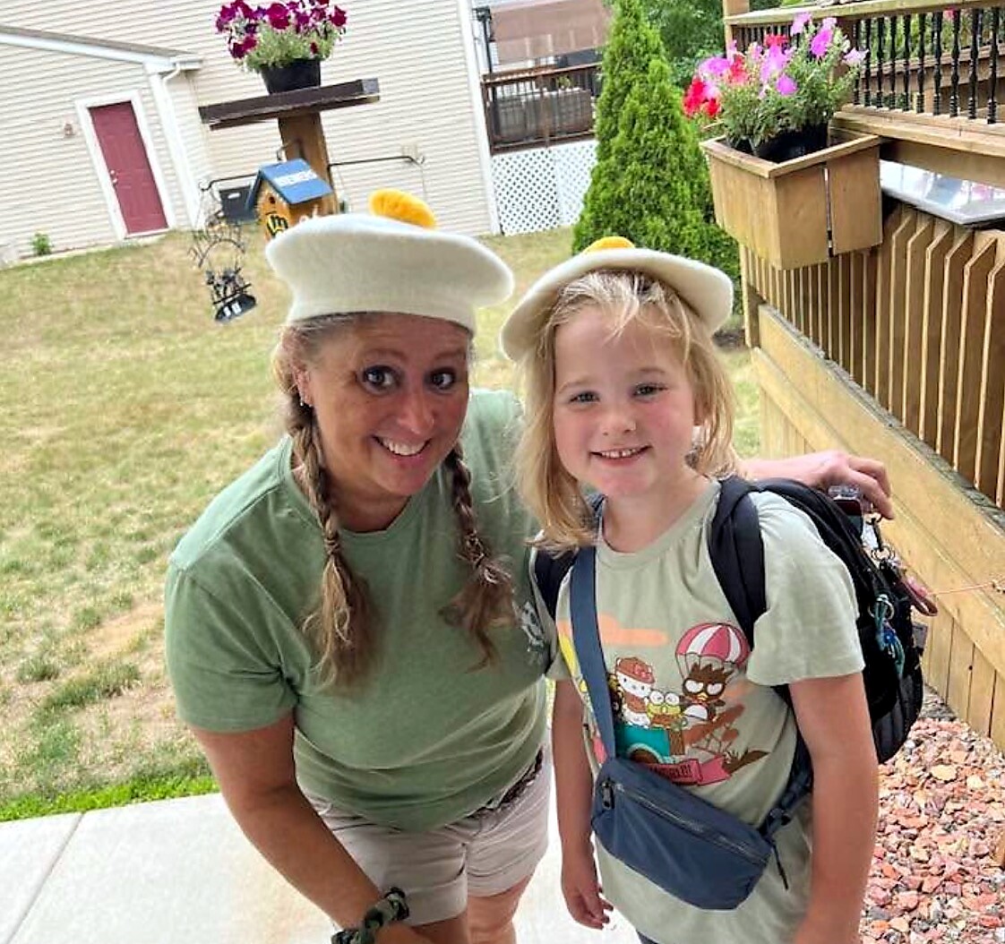 Woman and girl wearing white chef hats pose on a backyard porch, both in green shirts.