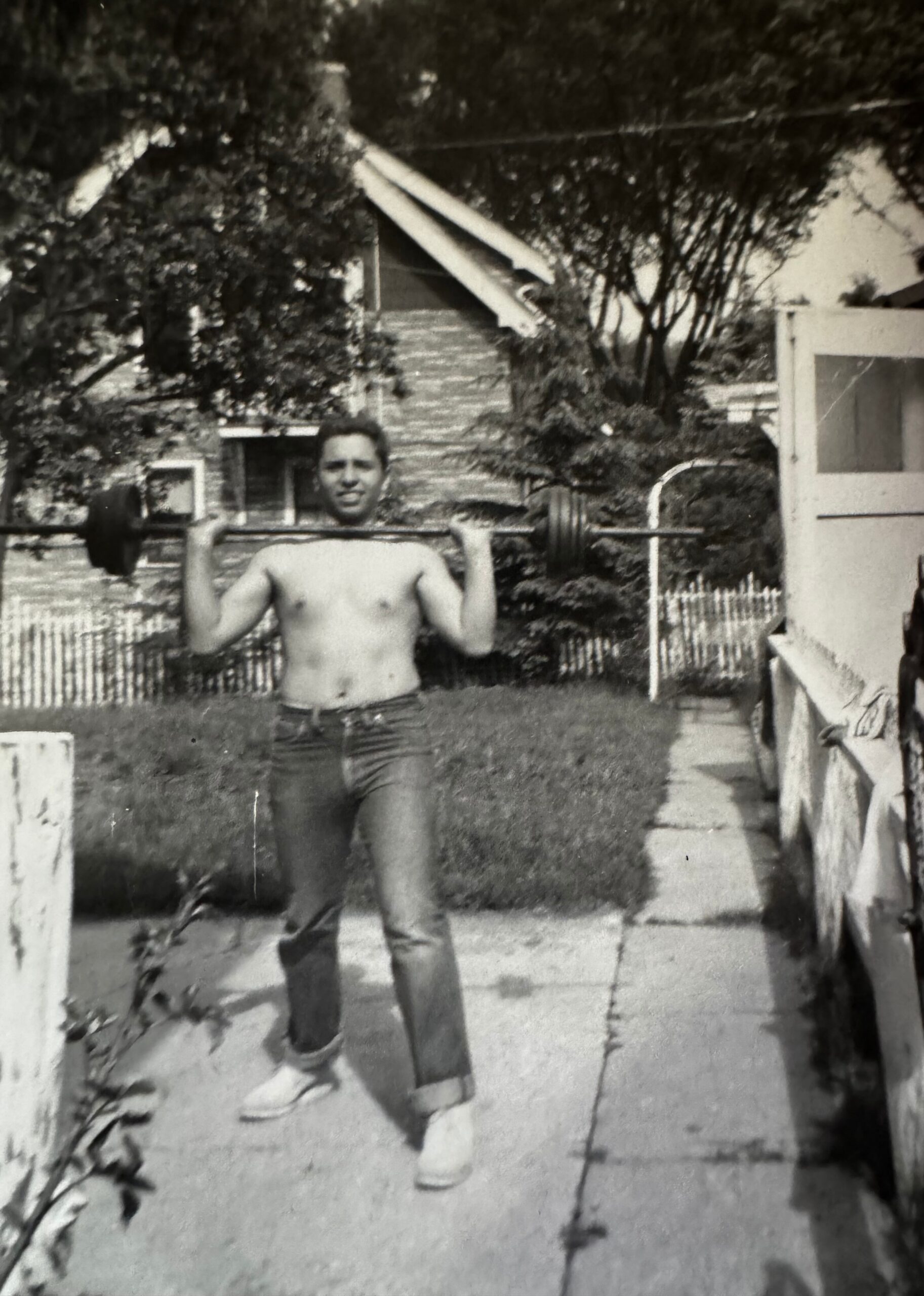Black-and-white photo of a shirtless man lifting a barbell in a fenced backyard with a house and stairs in the background.