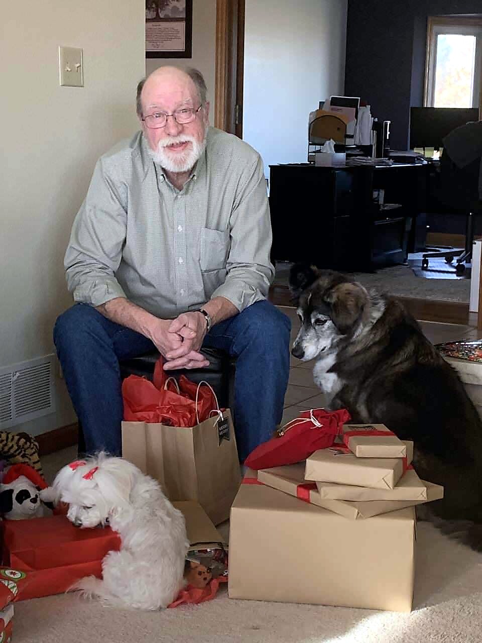 Older man with glasses sits on floor among wrapped gifts and two dogs in a home setting, holiday presents scattered around.