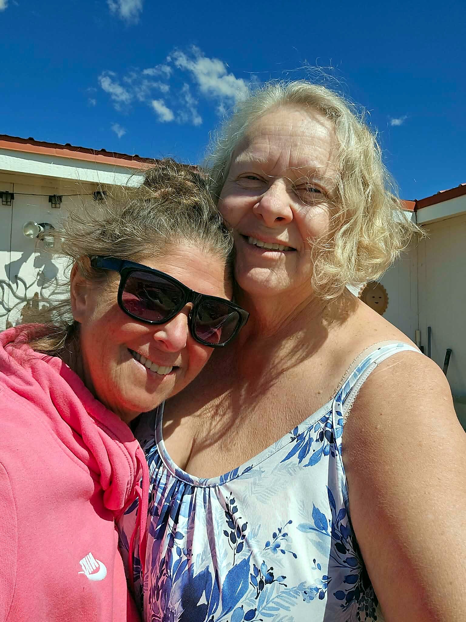 Two women smiling for a selfie outdoors, with a bright blue sky in the background.