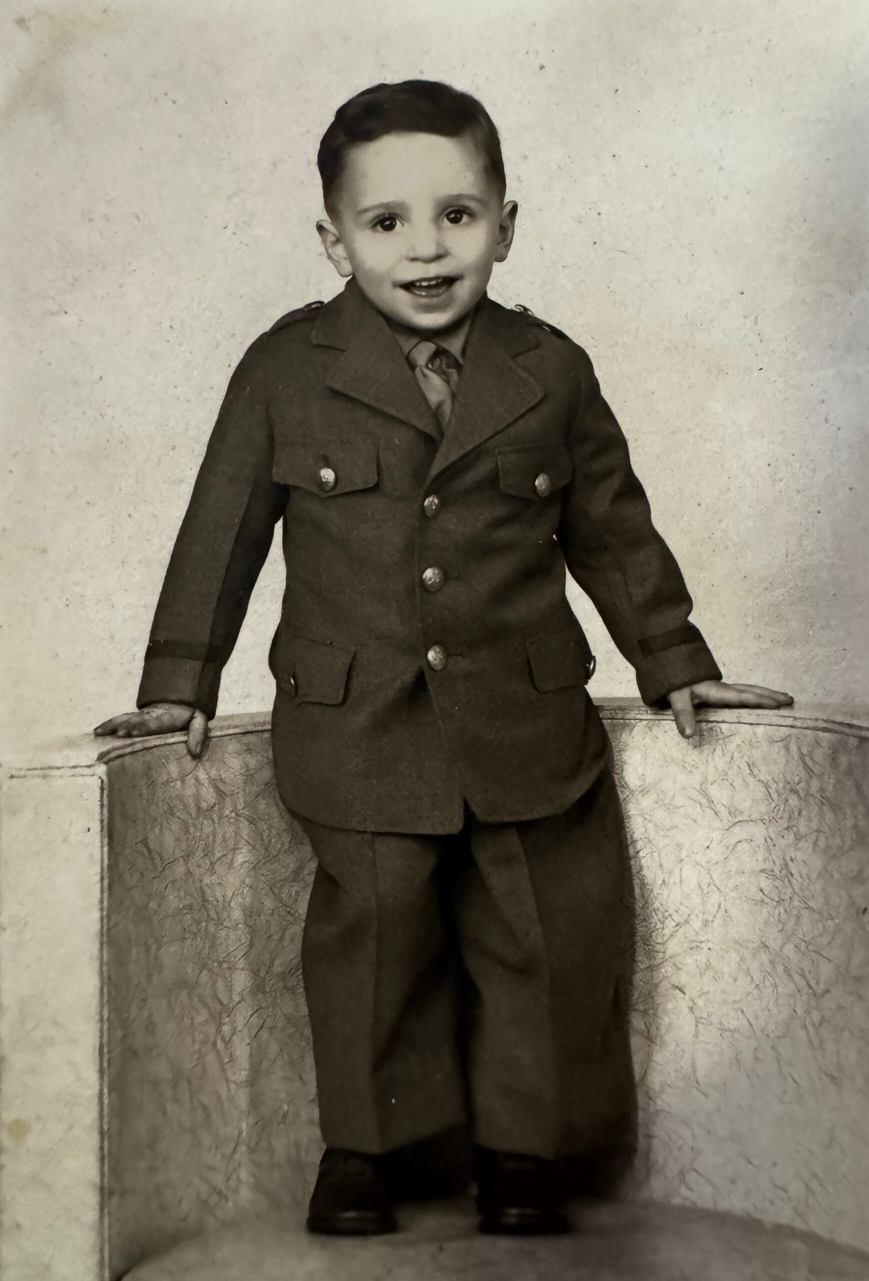 Young child wearing a dark military-style jacket with brass buttons, standing and smiling for a formal studio portrait.