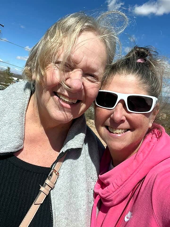 Two smiling women posing for a selfie outdoors on a sunny day; one with light hair in a gray jacket, the other in a pink hoodie and white sunglasses.