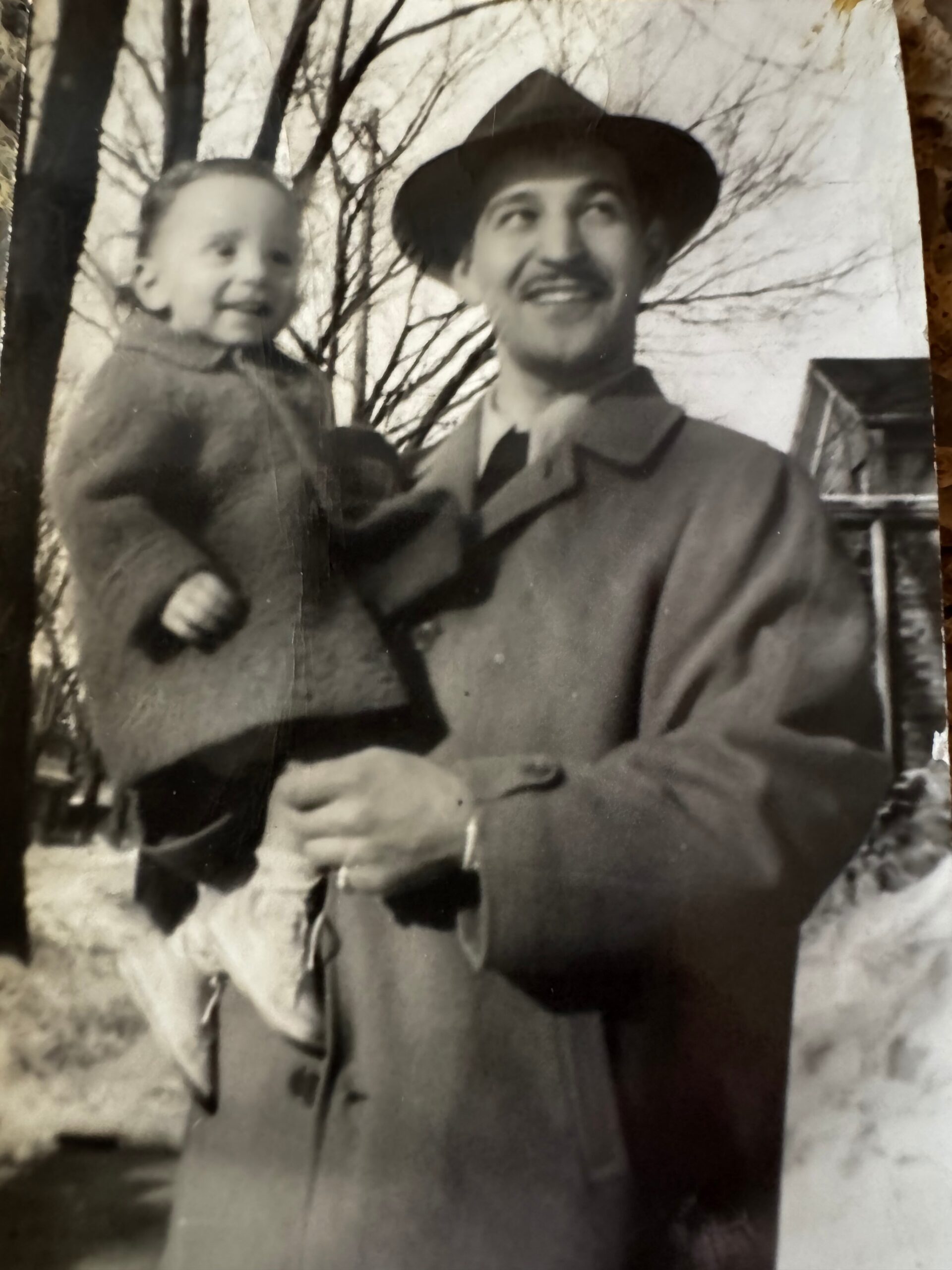 Man wearing a hat and coat, smiling, carries a young child outdoors with bare trees in the background.