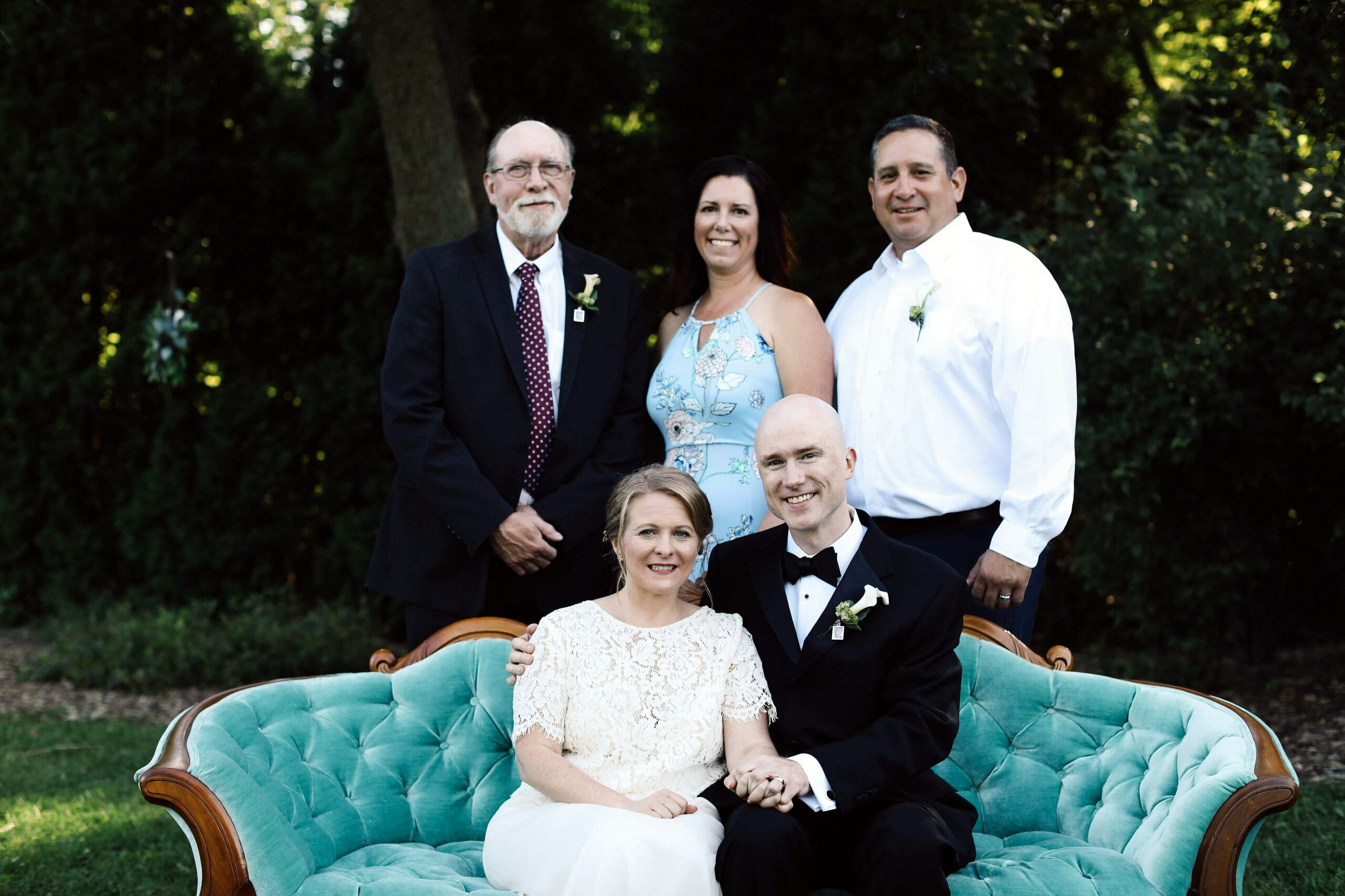 Wedding party portrait outdoors: a couple seated on a teal tufted sofa, with guests in formal attire standing behind them, greenery in the background.