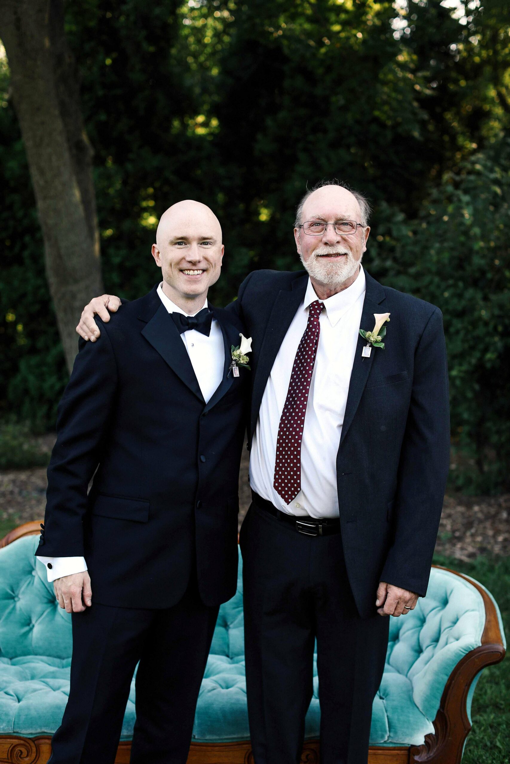 Two men in formal suits pose together outdoors, one younger bald man with a bow tie and an older man with glasses and a boutonniere, smiling.