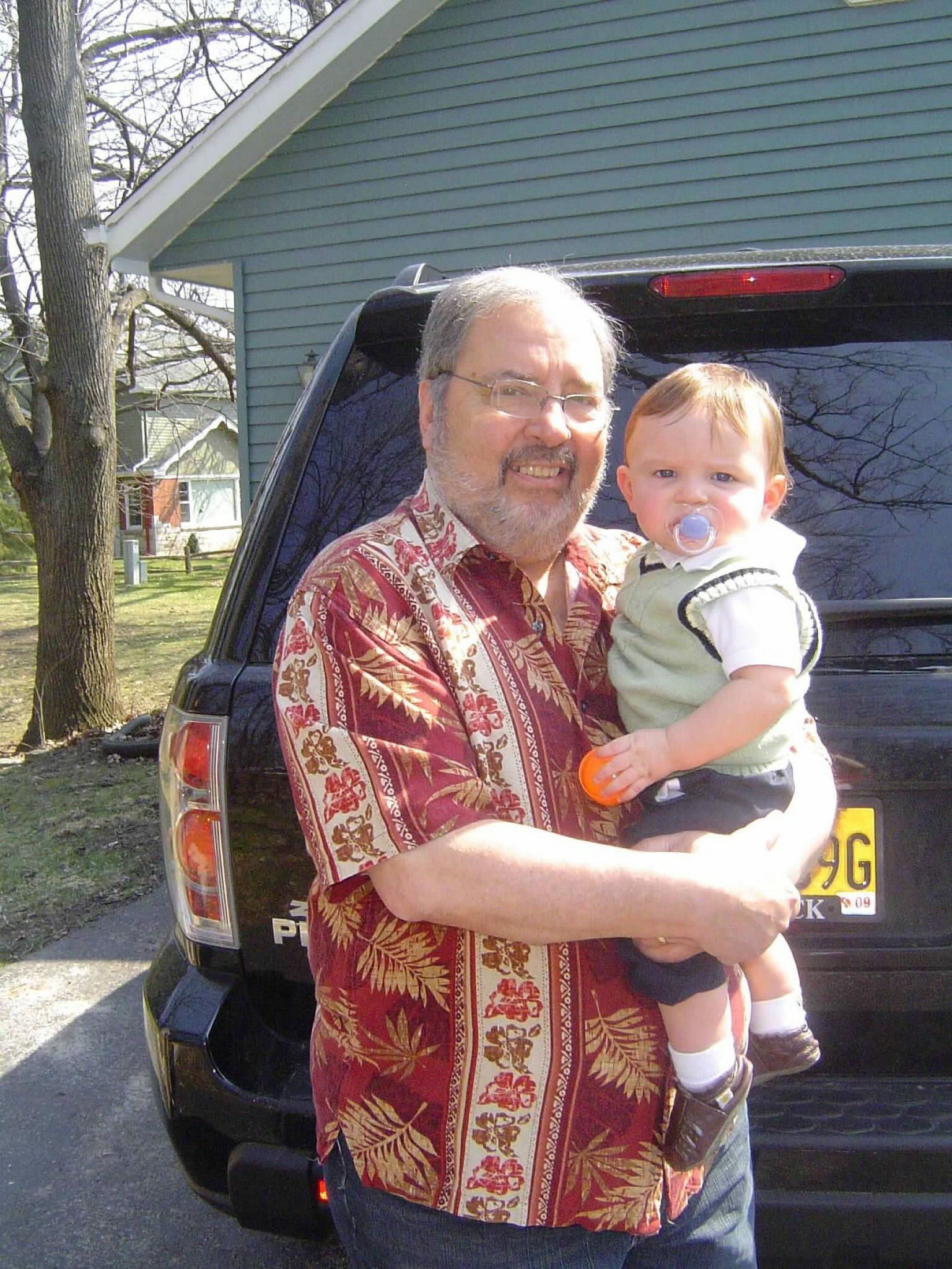 An older man in a red patterned shirt holds a baby with a pacifier, standing in front of a dark SUV outdoors.