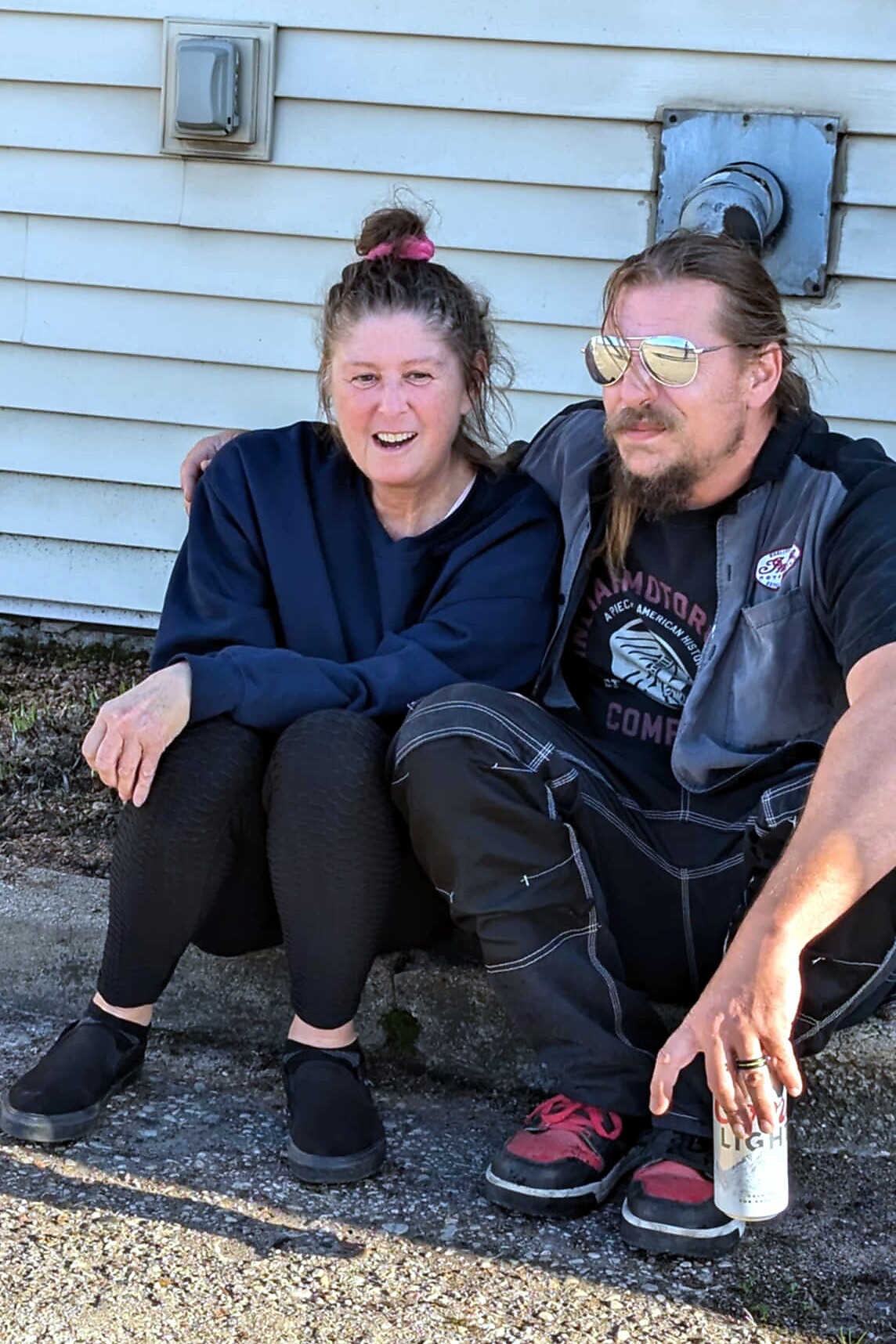 Woman and man sit close together on a curb against a light-blue house, smiling at the camera; the man wears sunglasses and holds a beer can.