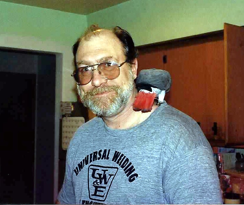 Older man with glasses and gray beard wearing a gray 'Universal Wiring' T-shirt, standing indoors with a red and black object tucked over his shoulder.