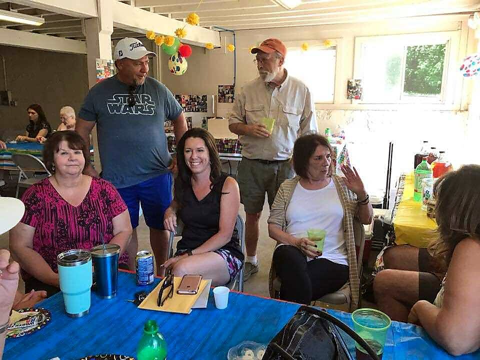 Group of adults at an indoor casual gathering, seated around a blue table with drinks and snacks, balloons in the background.